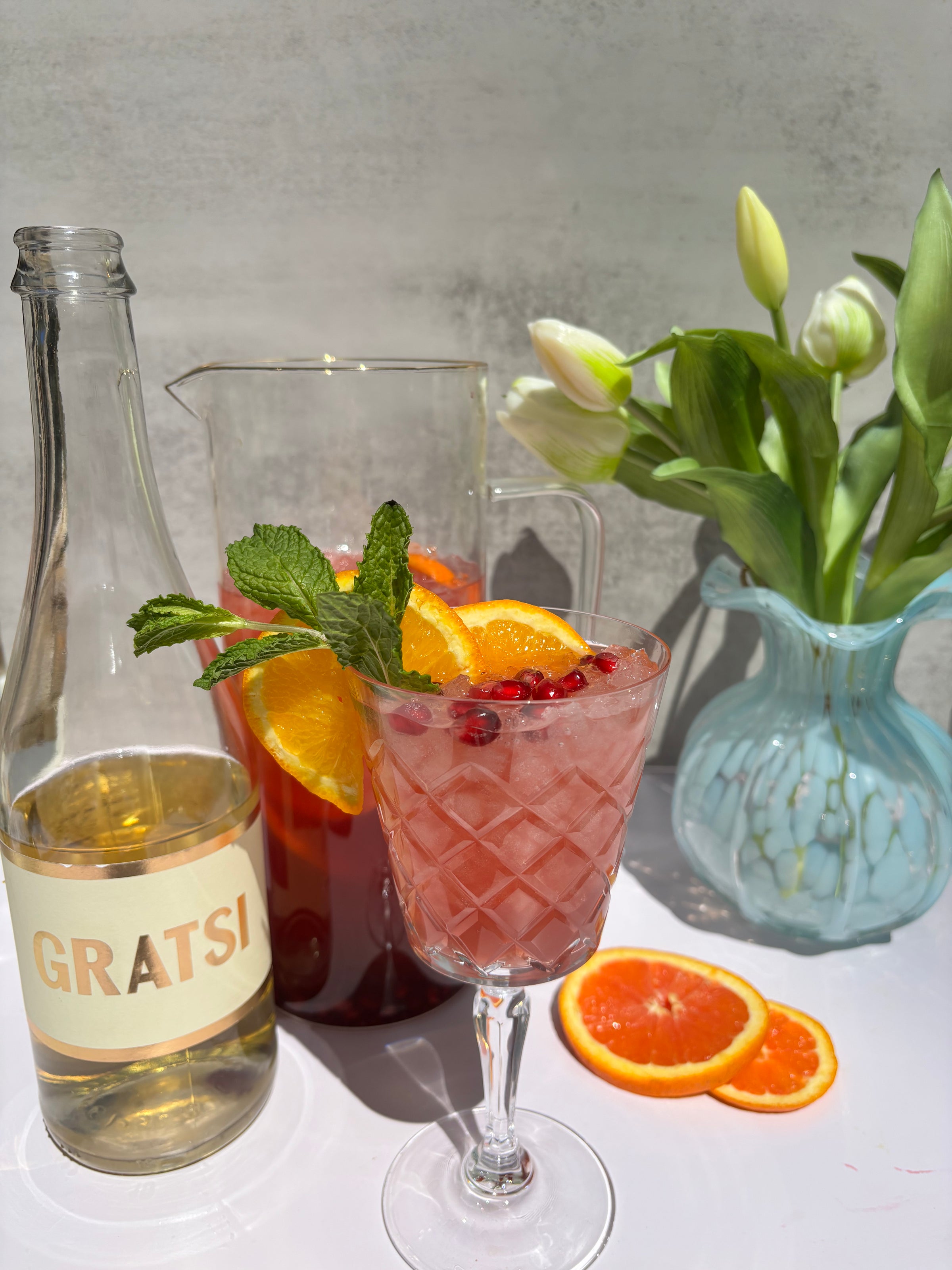 A refreshing cocktail garnished with orange slices, pomegranate seeds, and mint sits in front of a pitcher, a GRATSİ wine bottle, sliced oranges, and a blue vase with white tulips on a sunlit table.