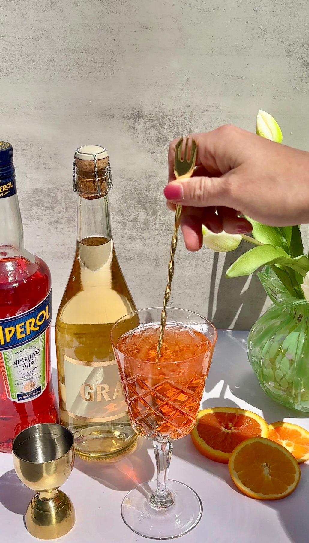 A hand stirs an orange cocktail in a glass, surrounded by Aperol, a bottle of sparkling wine, a jigger, orange slices, and a vase with white tulips on a sunlit surface.