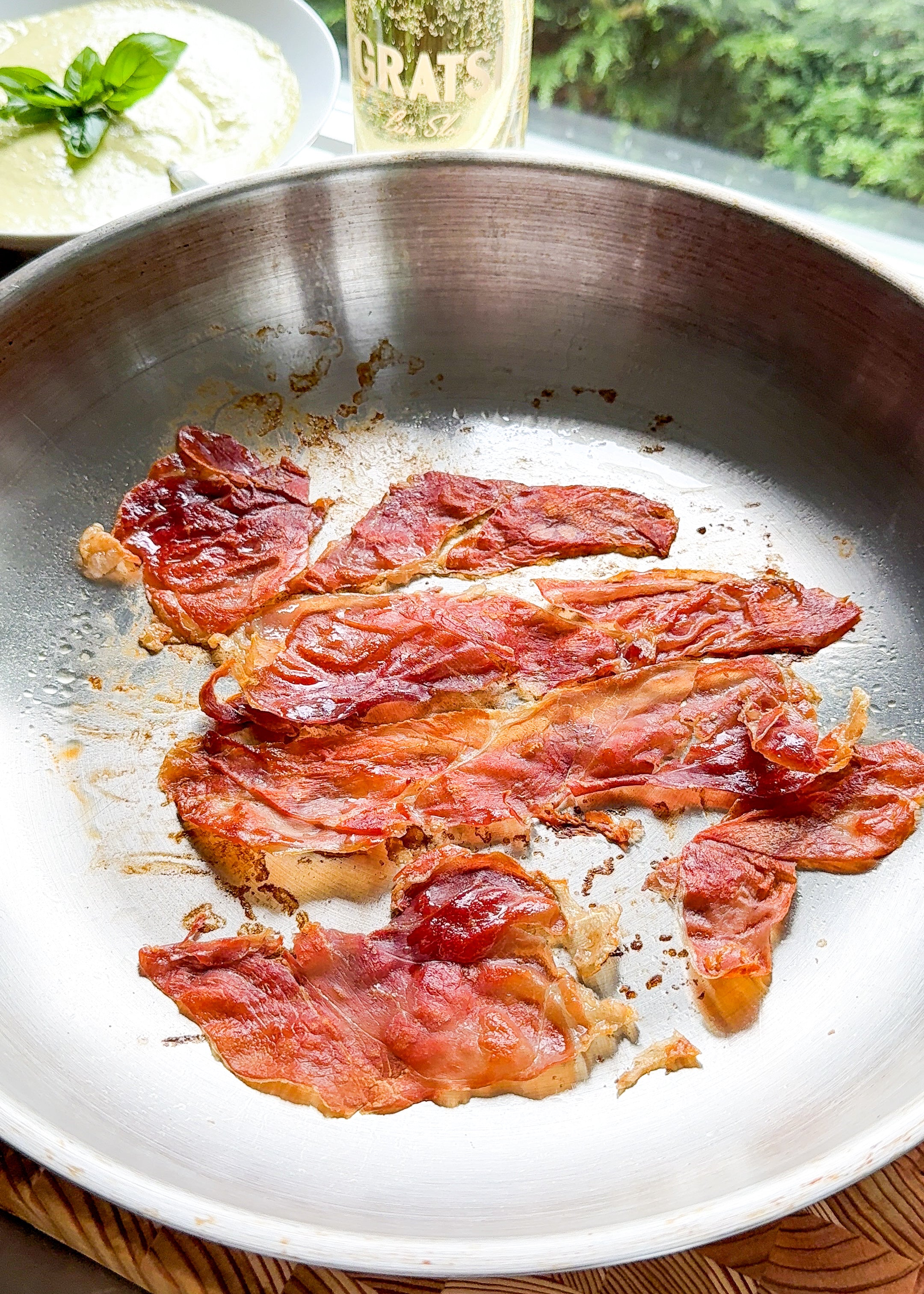 Several slices of crispy, cooked prosciutto lie in a silver pan on a wooden surface. In the background, there is a plate of food and a bottle, with greenery visible outside a window.