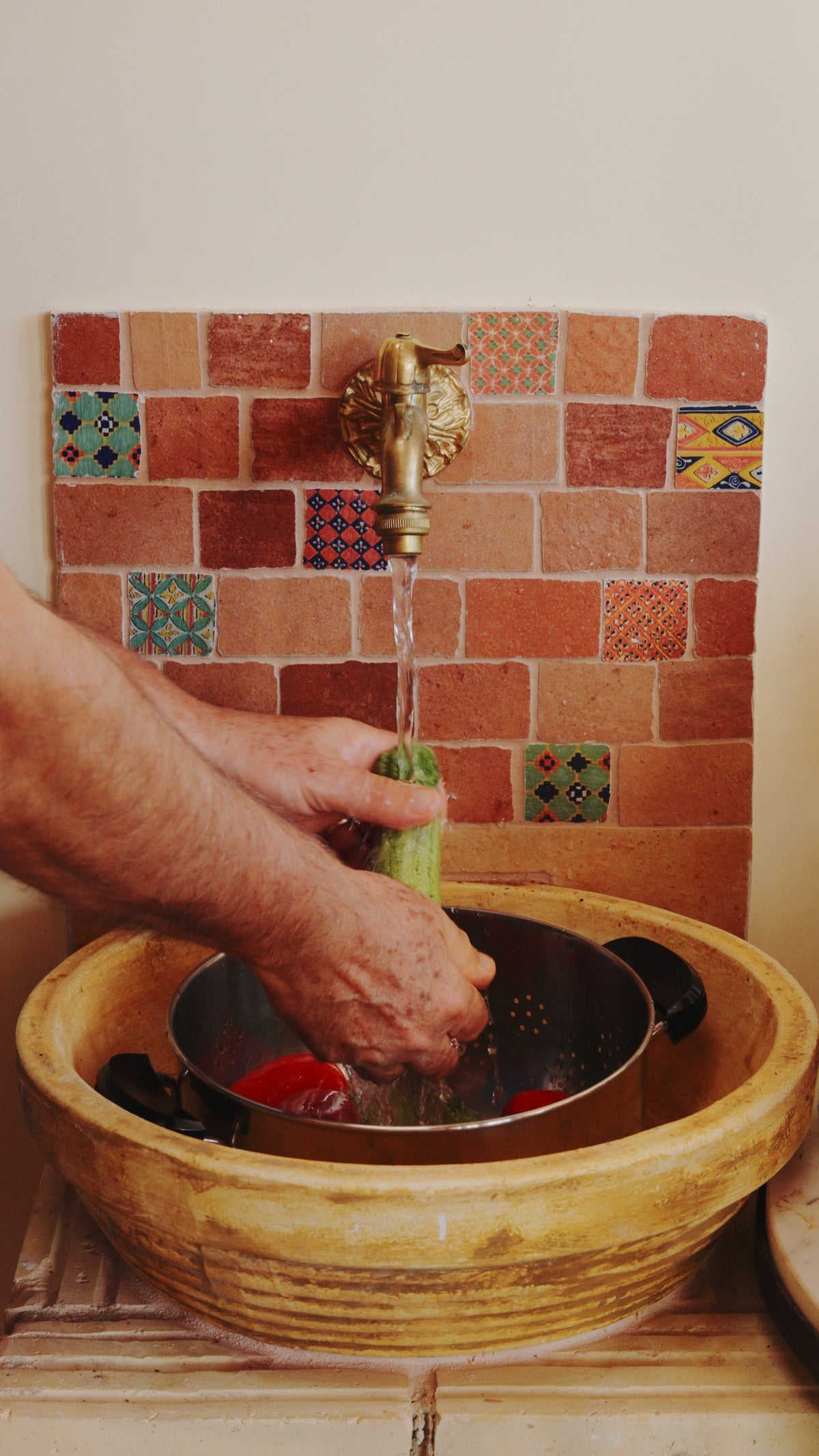 Hands washing a cucumber under a brass faucet mounted on a terracotta tiled wall, with a colander inside a large stone basin.