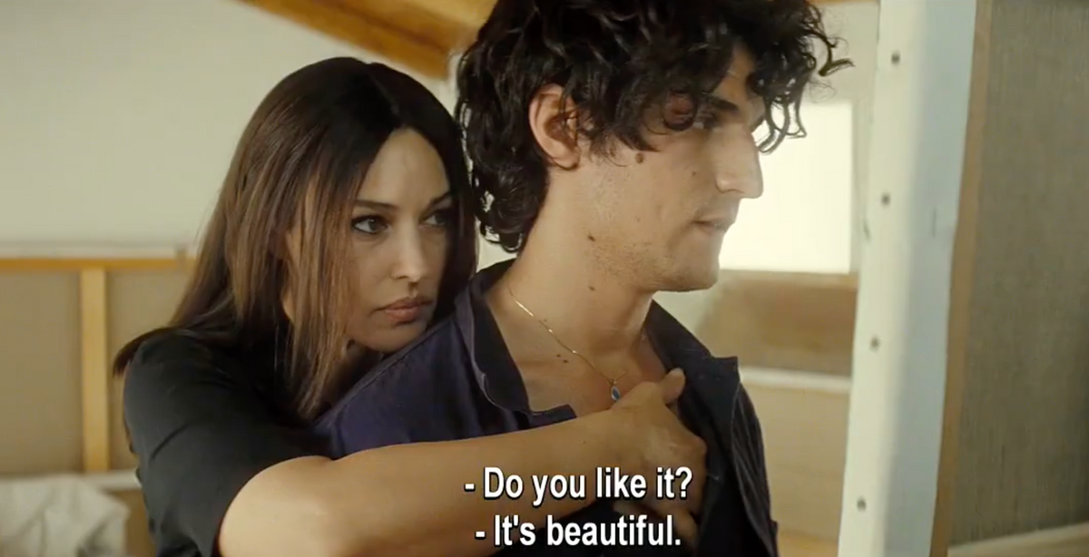 A woman stands behind a man, putting a necklace around his neck. She looks intent, while he looks ahead. The subtitles read: - Do you like it? - Its beautiful..