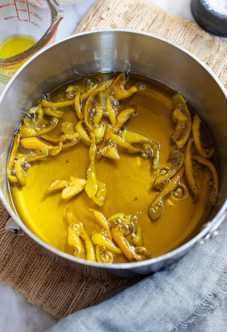 A stainless steel pot filled with olive oil and lemon peels sits on a burlap mat. A glass measuring cup with yellow liquid is nearby, and a gray cloth is partially visible at the bottom right.
