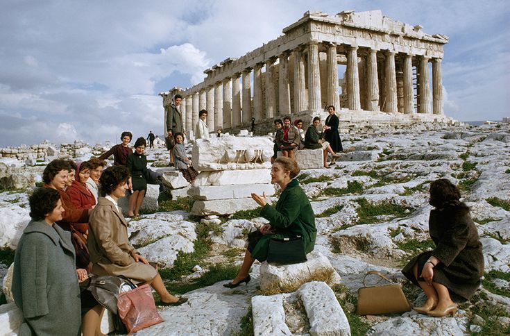 A group of women sit and stand on rocky ground near the Parthenon in Athens, Greece, listening to a woman in green who appears to be speaking or guiding them. The ancient temple stands majestically in the background.