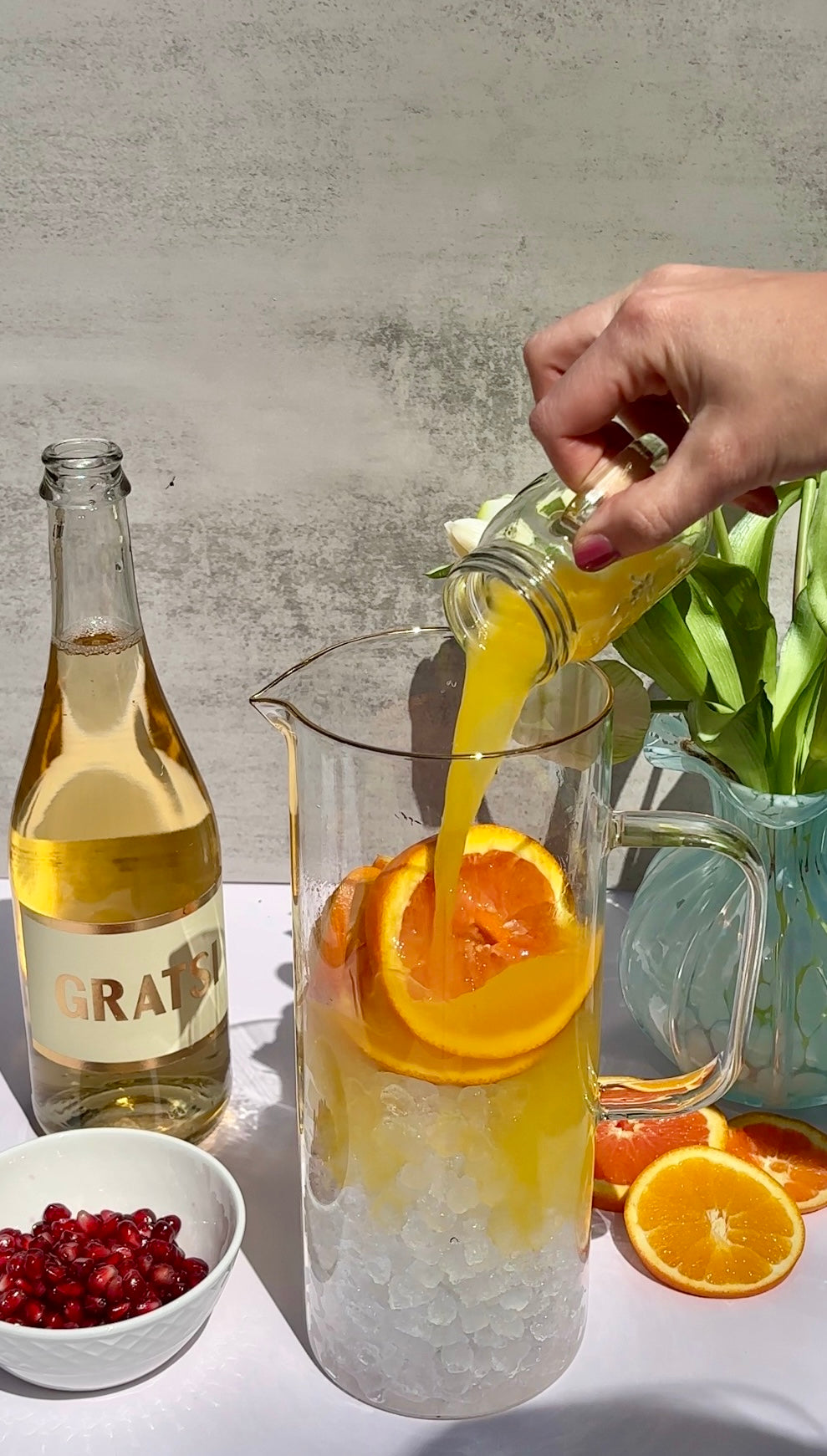 A hand pours orange juice from a small glass jar into a pitcher filled with ice and orange slices. Nearby are a bottle labeled “Gratsi,” a bowl of pomegranate seeds, and a vase with flowers.