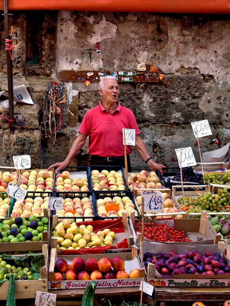 An elderly man in a pink shirt stands behind a vibrant fruit stall at an outdoor market, surrounded by peaches, grapes, figs, and other fresh produce, with handwritten price signs on display.