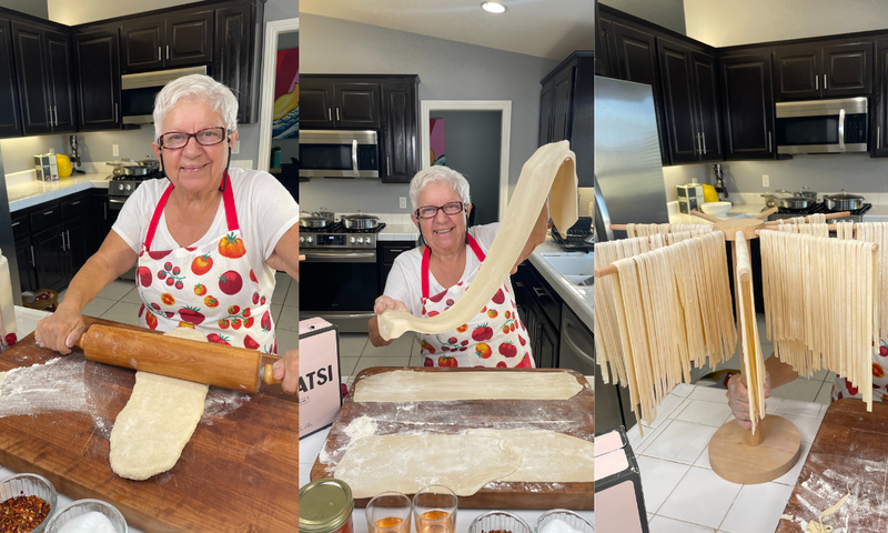 A smiling older woman in a kitchen rolls, stretches, and hangs homemade pasta dough to dry, wearing a red apron with tomatoes and cherries. The kitchen has dark cabinets and modern appliances.