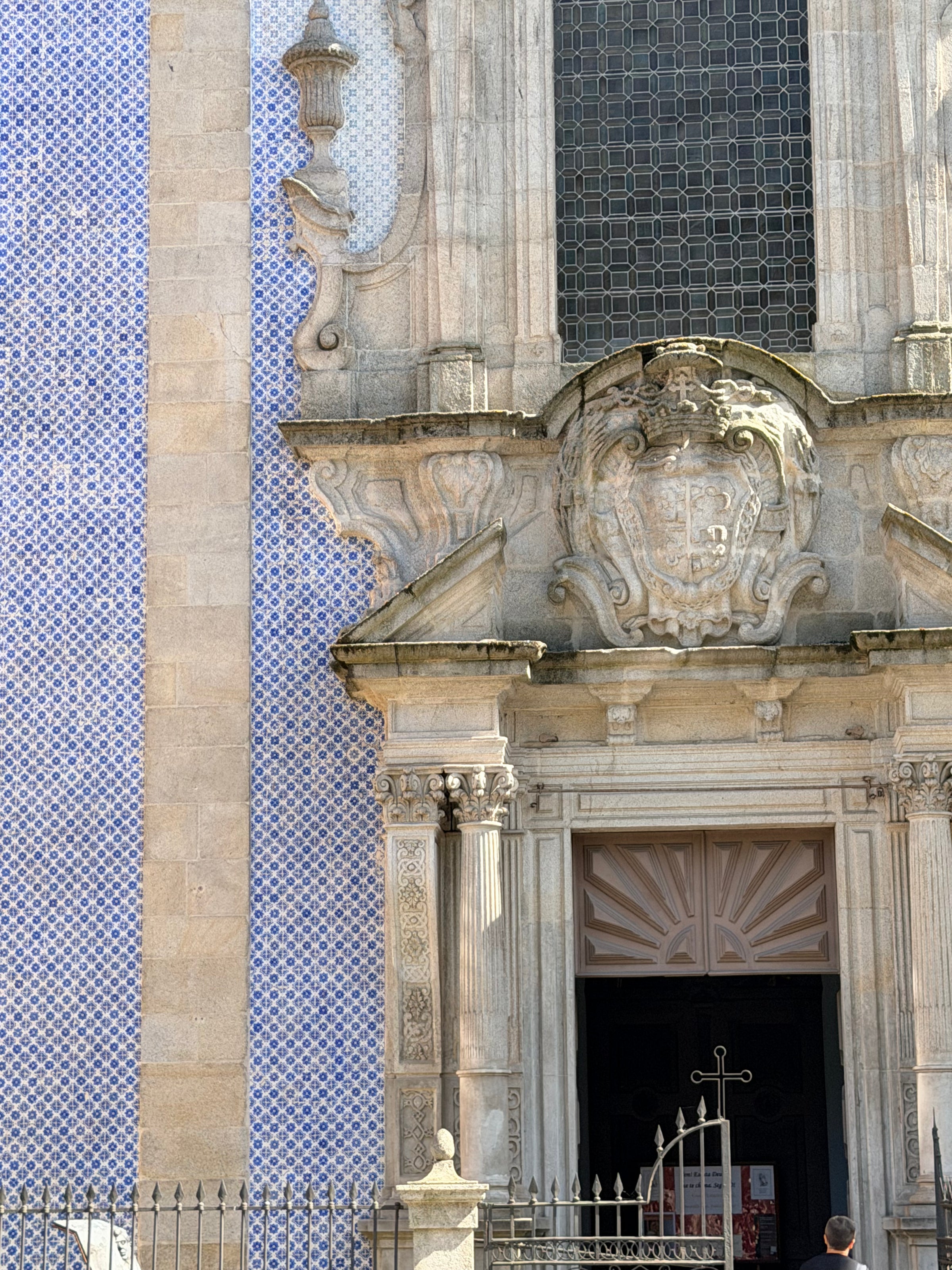 Stone entrance of a historic building with ornate carvings, a large coat of arms above the door, blue-and-white patterned tiles on the wall, and a decorative iron gate in front.