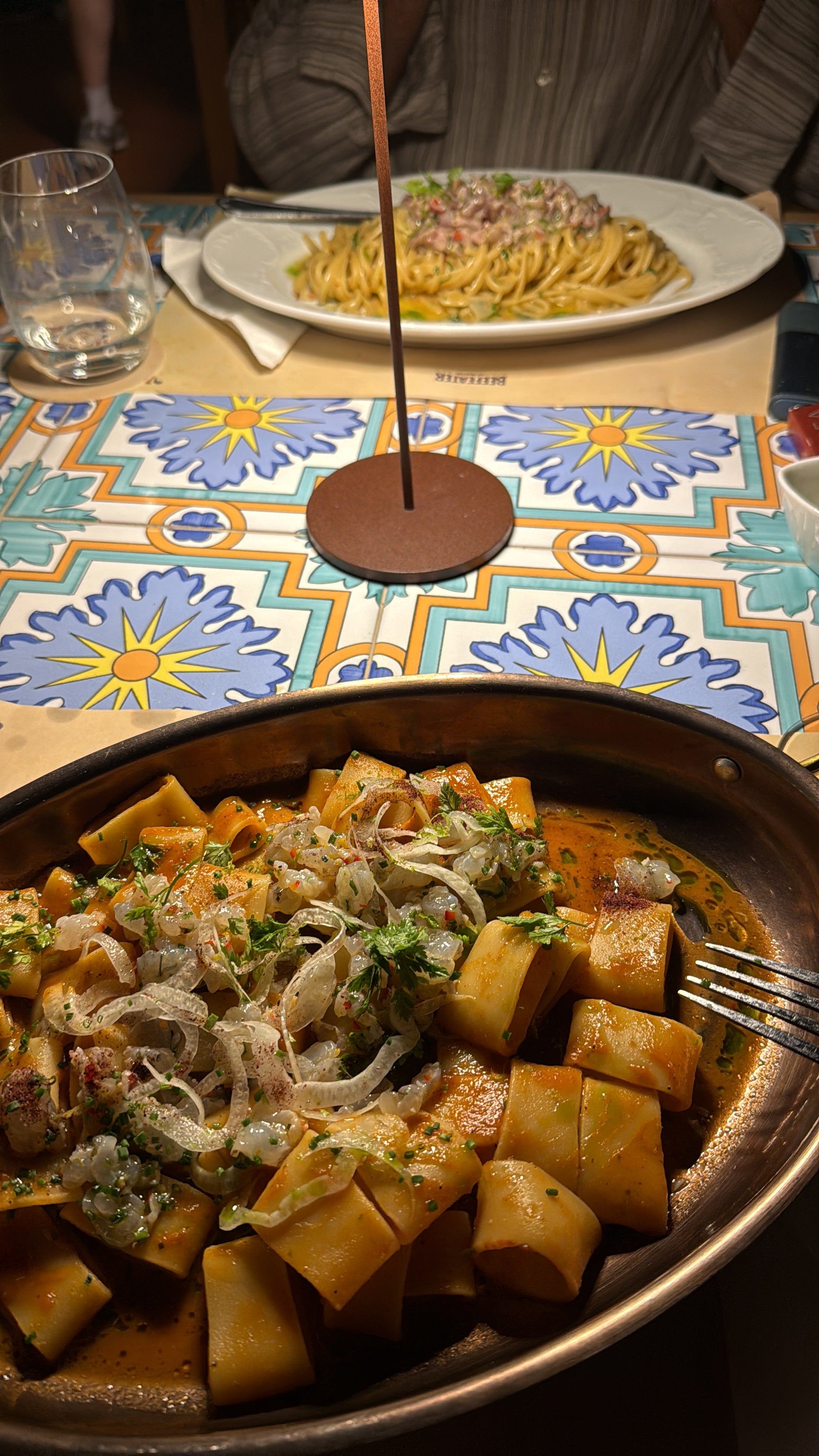 A dish of pasta with sauce and herbs in the foreground, another pasta plate in the background, on a colorful patterned table, with glasses and utensils visible.
