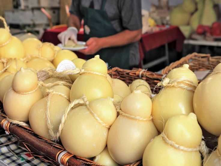 A basket filled with round, yellow cheese wheels tied with twine is displayed at a market stall. In the background, a person wearing an apron and gloves is slicing cheese samples.