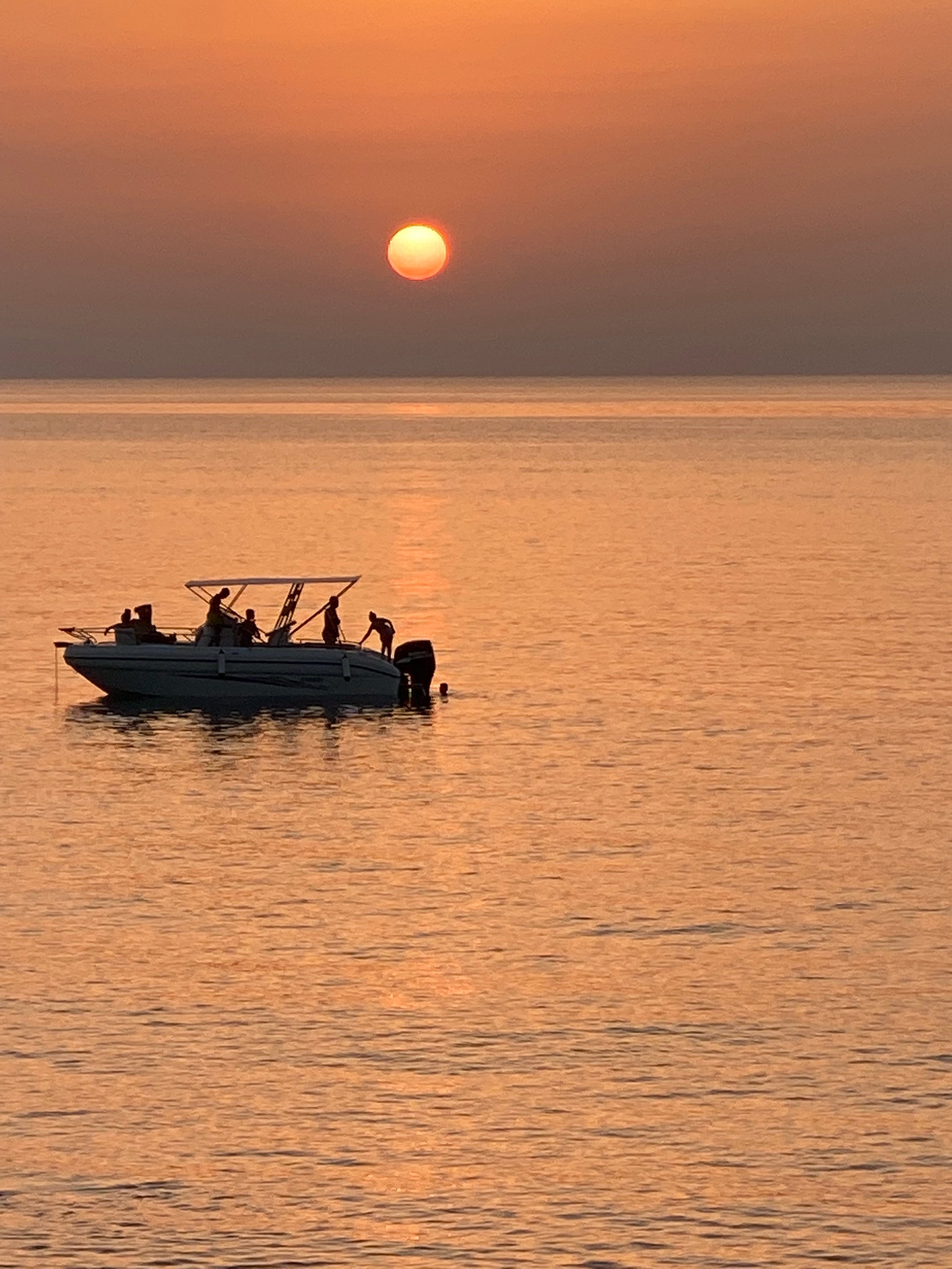 A small boat with several people on board is floating on calm water during sunset, with the orange sun low in the sky and its reflection shimmering on the sea.