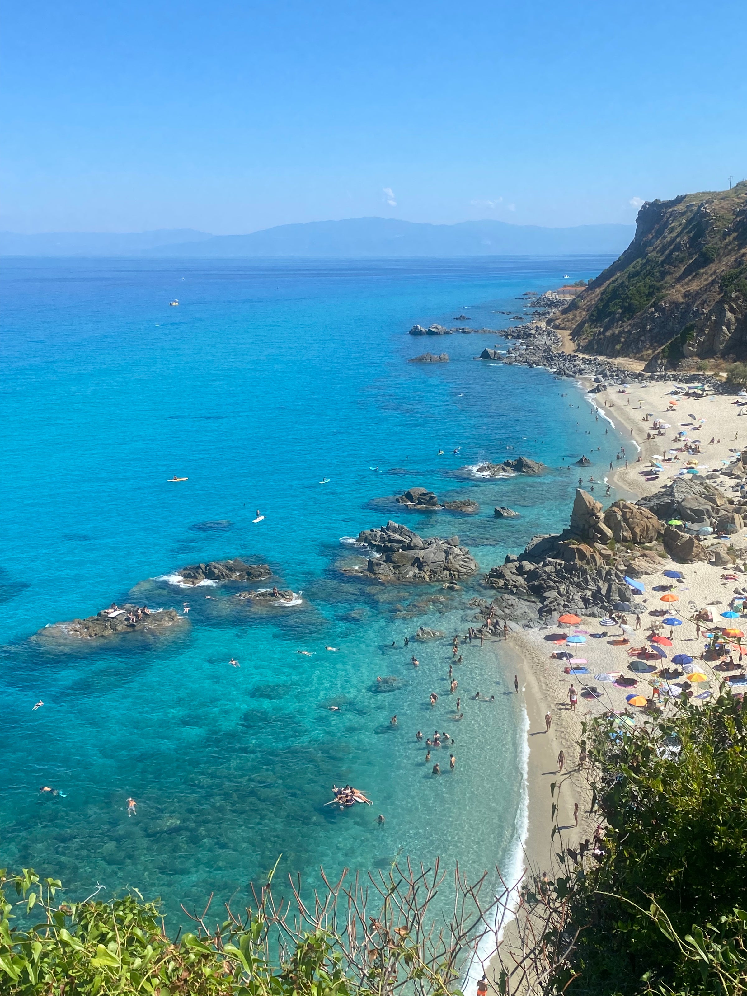 A scenic view of a turquoise sea with rocky outcrops and a sandy beach, where many people are swimming and sunbathing under colorful umbrellas. Hills and cliffs rise along the coastline under a clear blue sky.