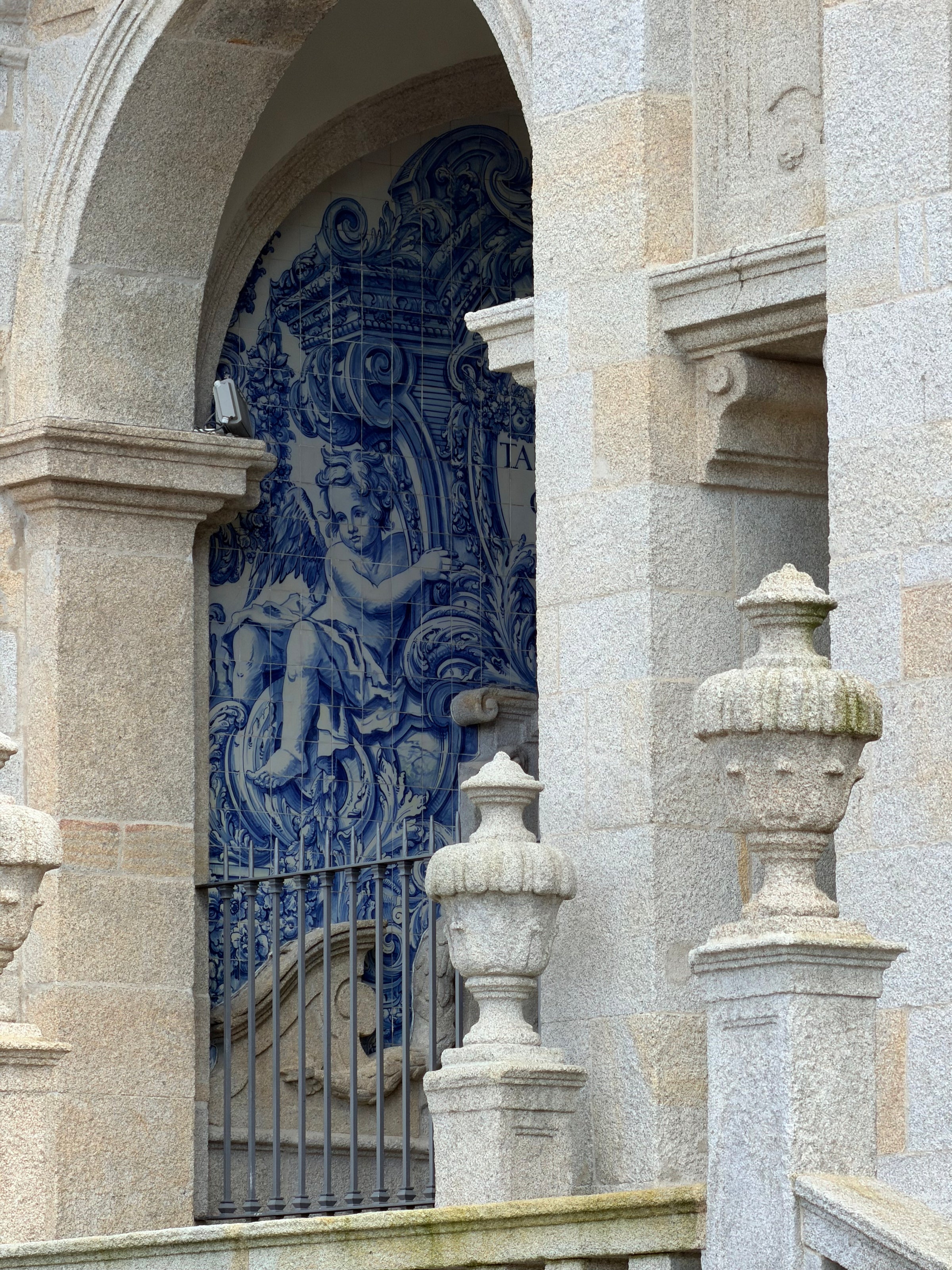 A detailed blue and white tile mural depicting a classical figure is framed by a stone archway and columns, with ornate stone balusters in the foreground.