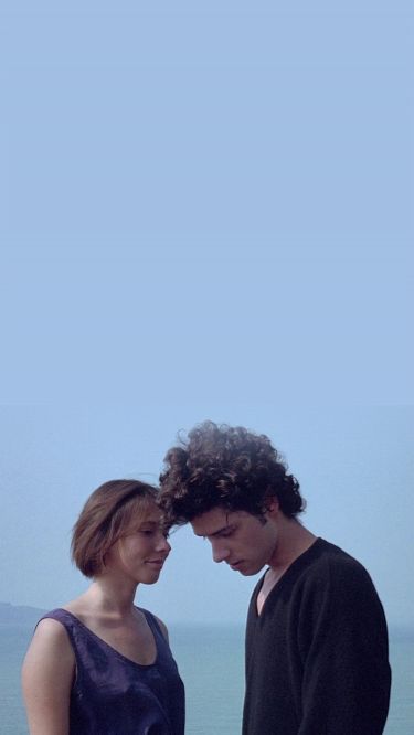 A young woman and man stand close together by the sea under a clear blue sky. The woman smiles gently while the man looks down, both appearing thoughtful and calm.