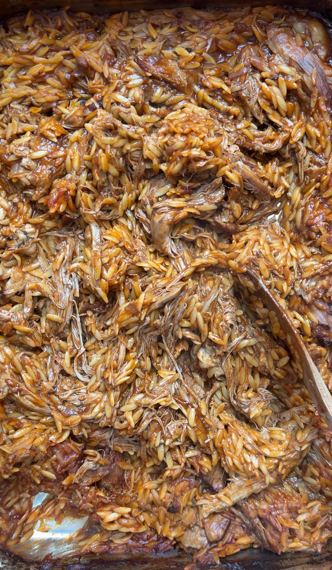 A close-up of a baked dish with shredded meat and orzo pasta mixed in a rich, brown sauce, with a wooden spoon stirring the mixture.