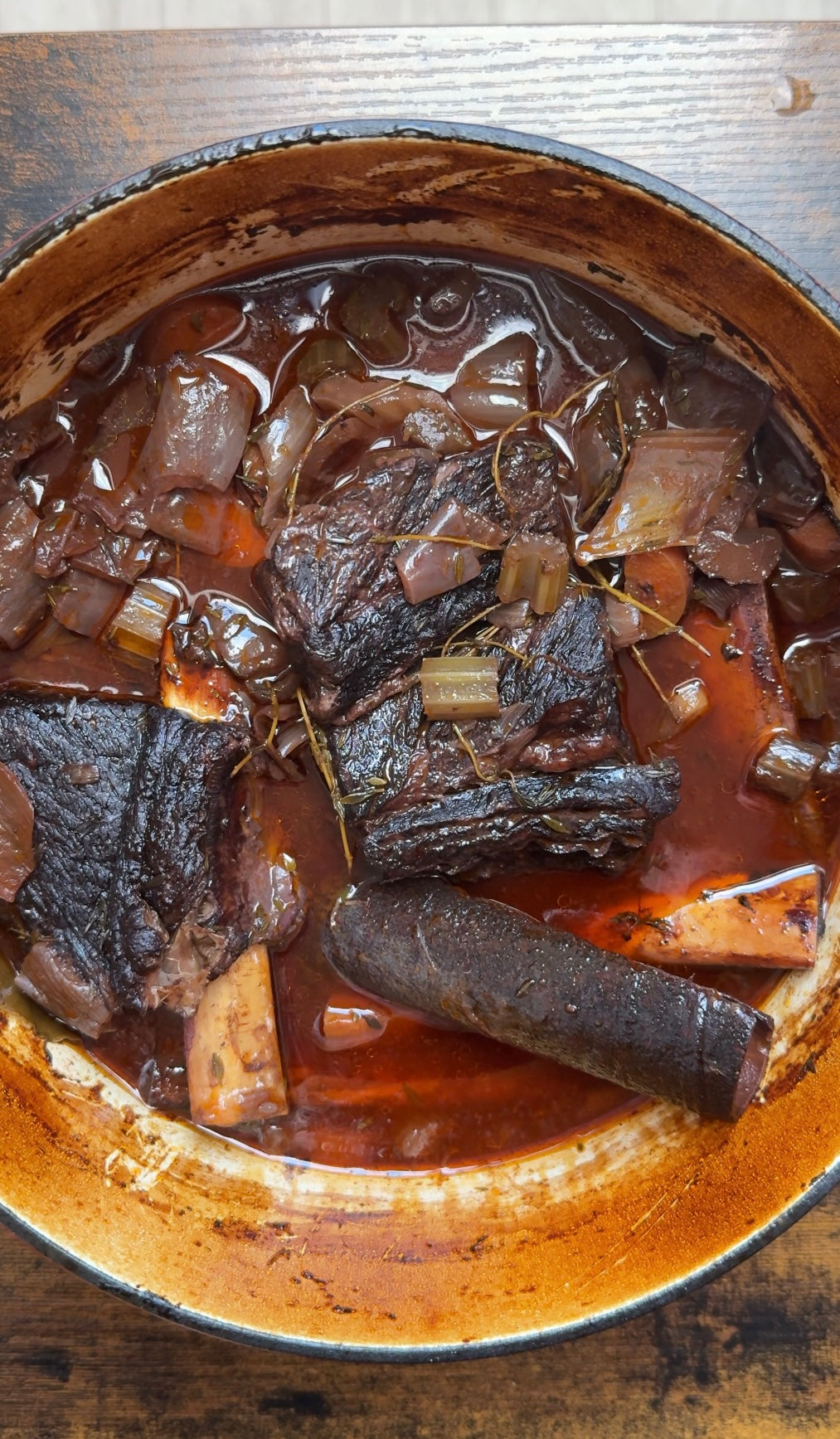 Braised short ribs in a Dutch oven with a rich, dark sauce, chunks of onion, celery, and herbs visible in the mix; the dish appears tender and cooked with a deep, flavorful glaze.