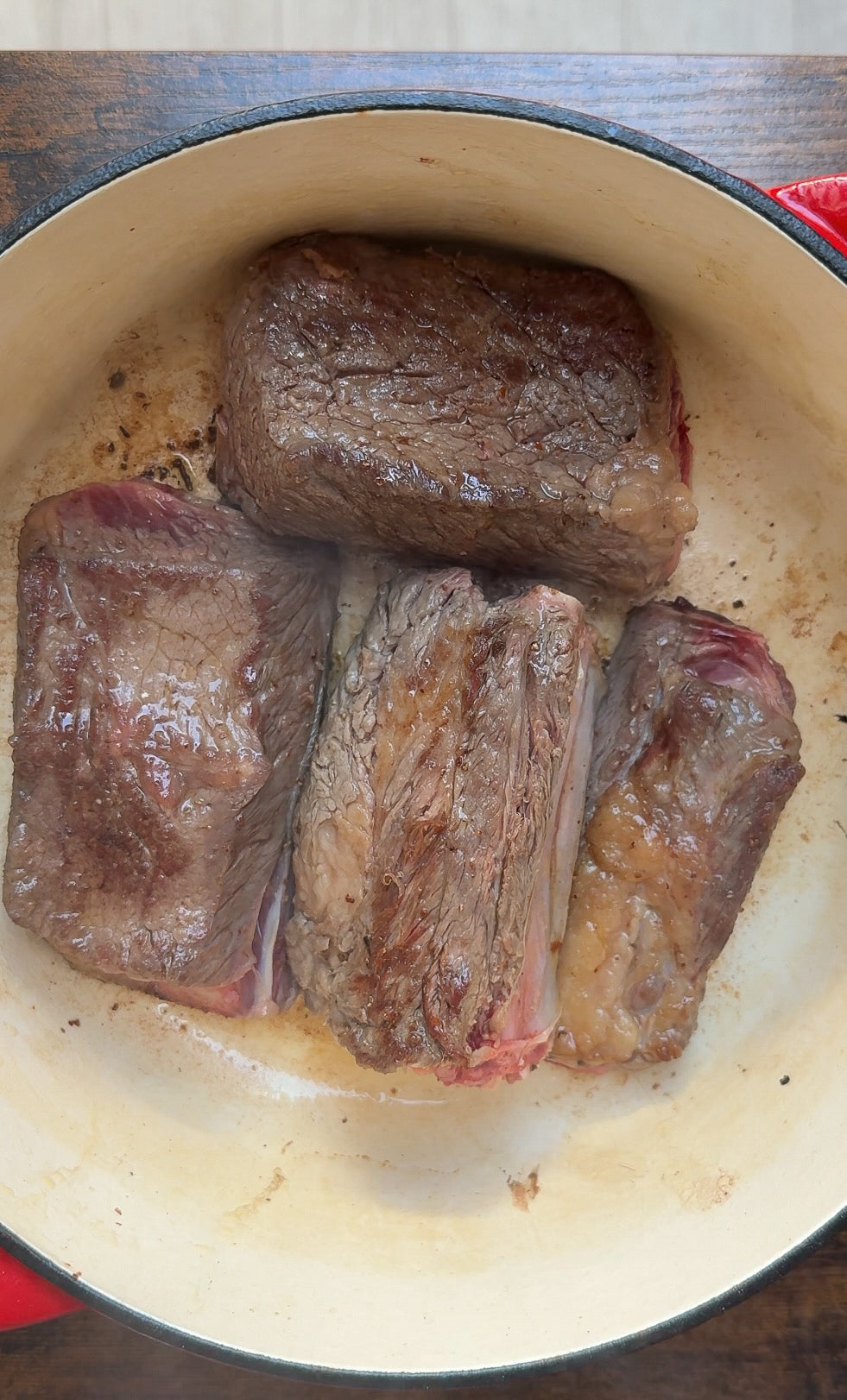 Four pieces of browned beef are searing in a round, cream-colored Dutch oven with a red rim. The beef has a caramelized crust and is arranged closely together.