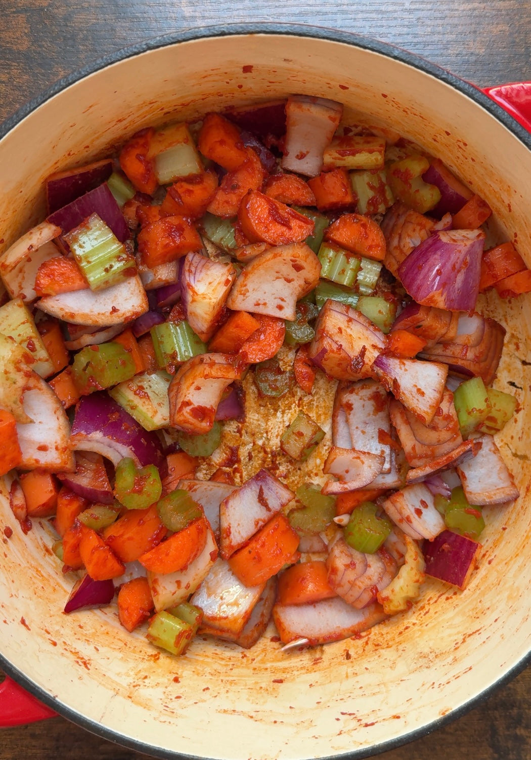 Chopped carrots, celery, and red onions seasoned with spices are sautéing in a red Dutch oven on a wooden surface.