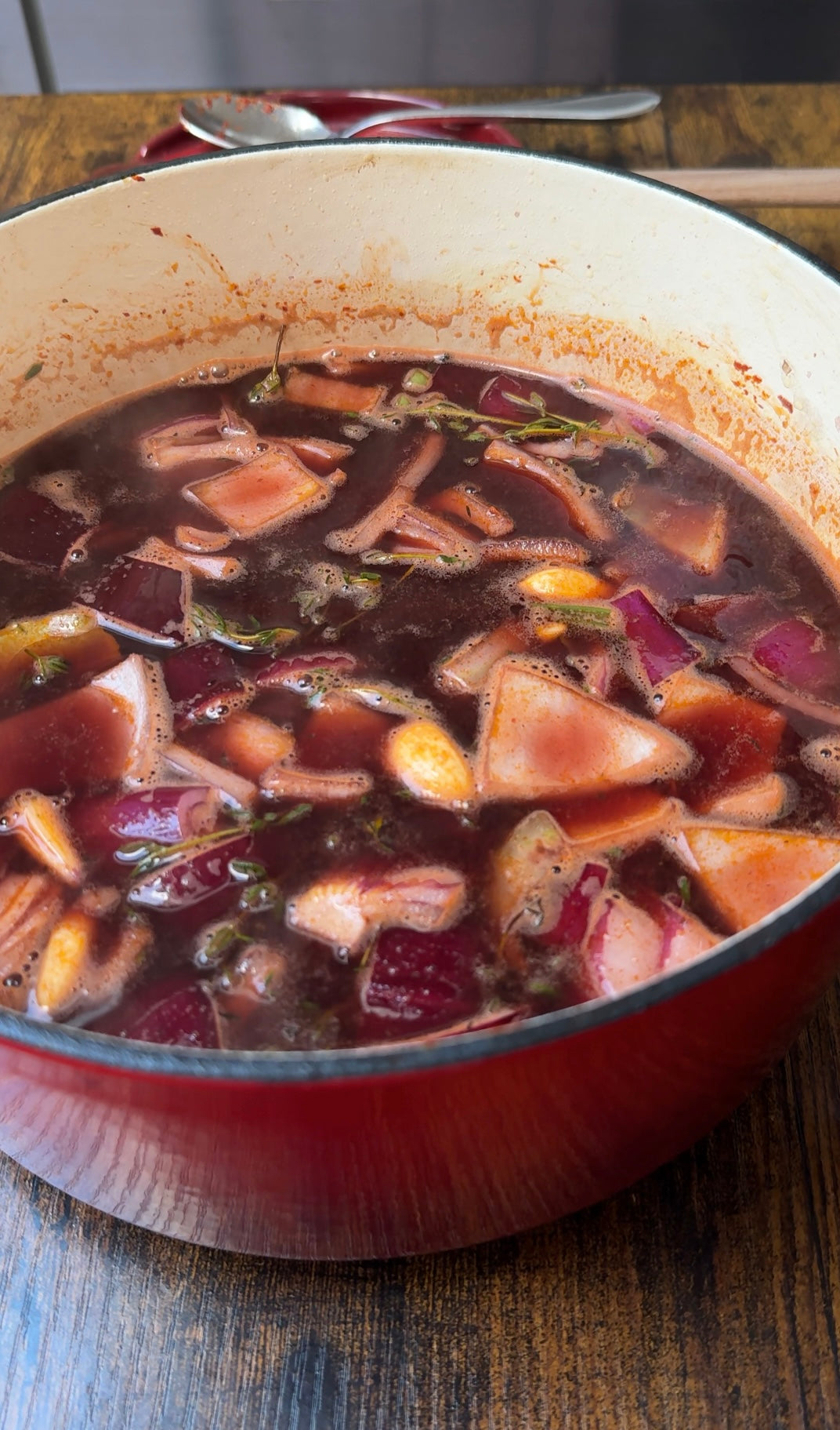 A red pot filled with a dark, broth-based soup containing chopped vegetables like carrots, onions, and beets, simmering on a wooden surface.
