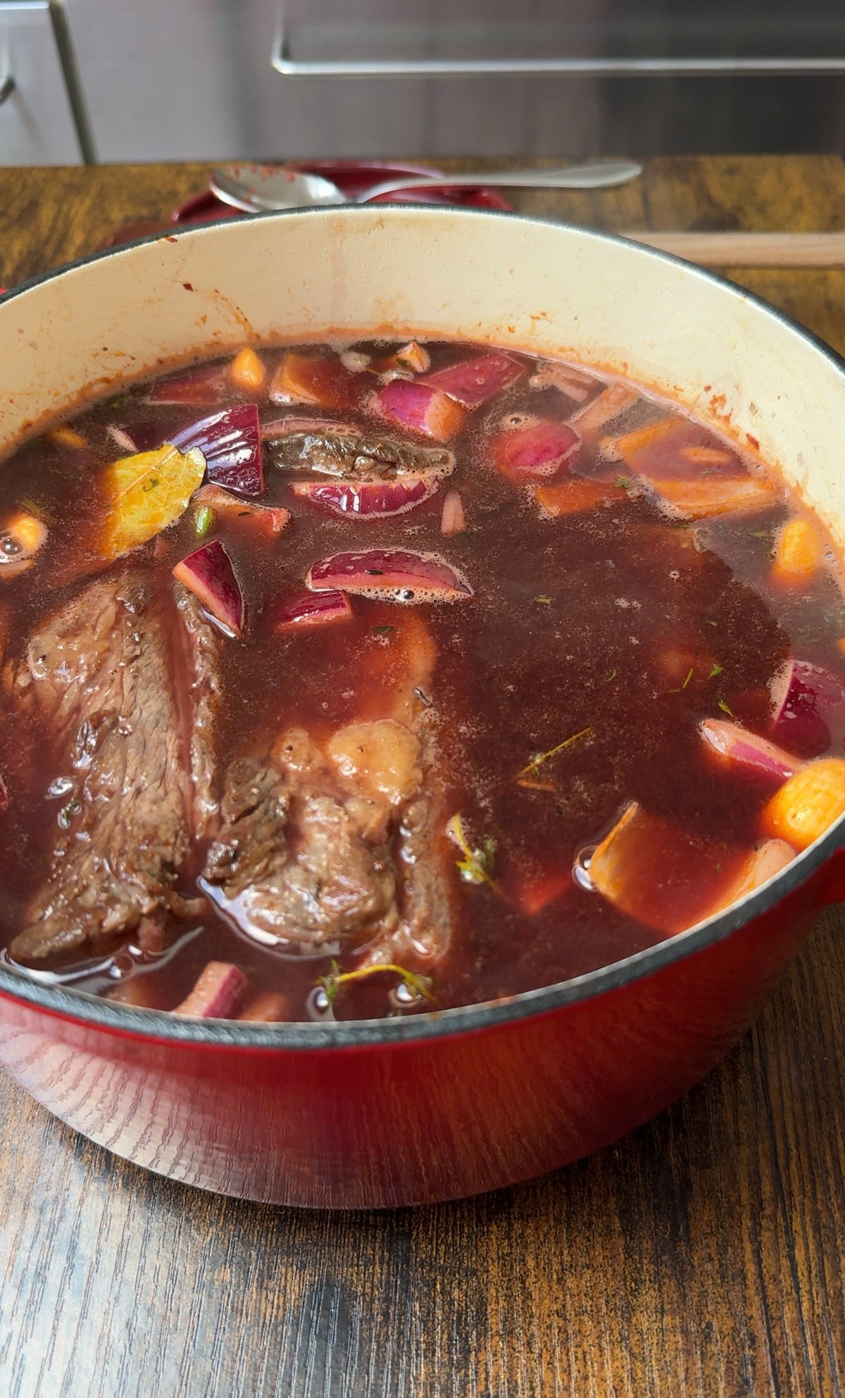 A red Dutch oven filled with beef stew, including chunks of beef, carrots, celery, and red onions in a rich, dark broth, sits on a wooden table.