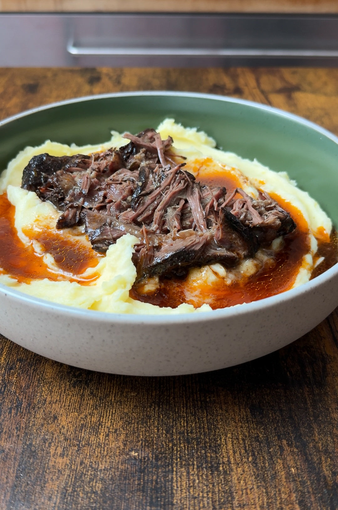 Shredded pot roast with rich brown gravy served over creamy mashed potatoes in a shallow bowl, set on a wooden table.
