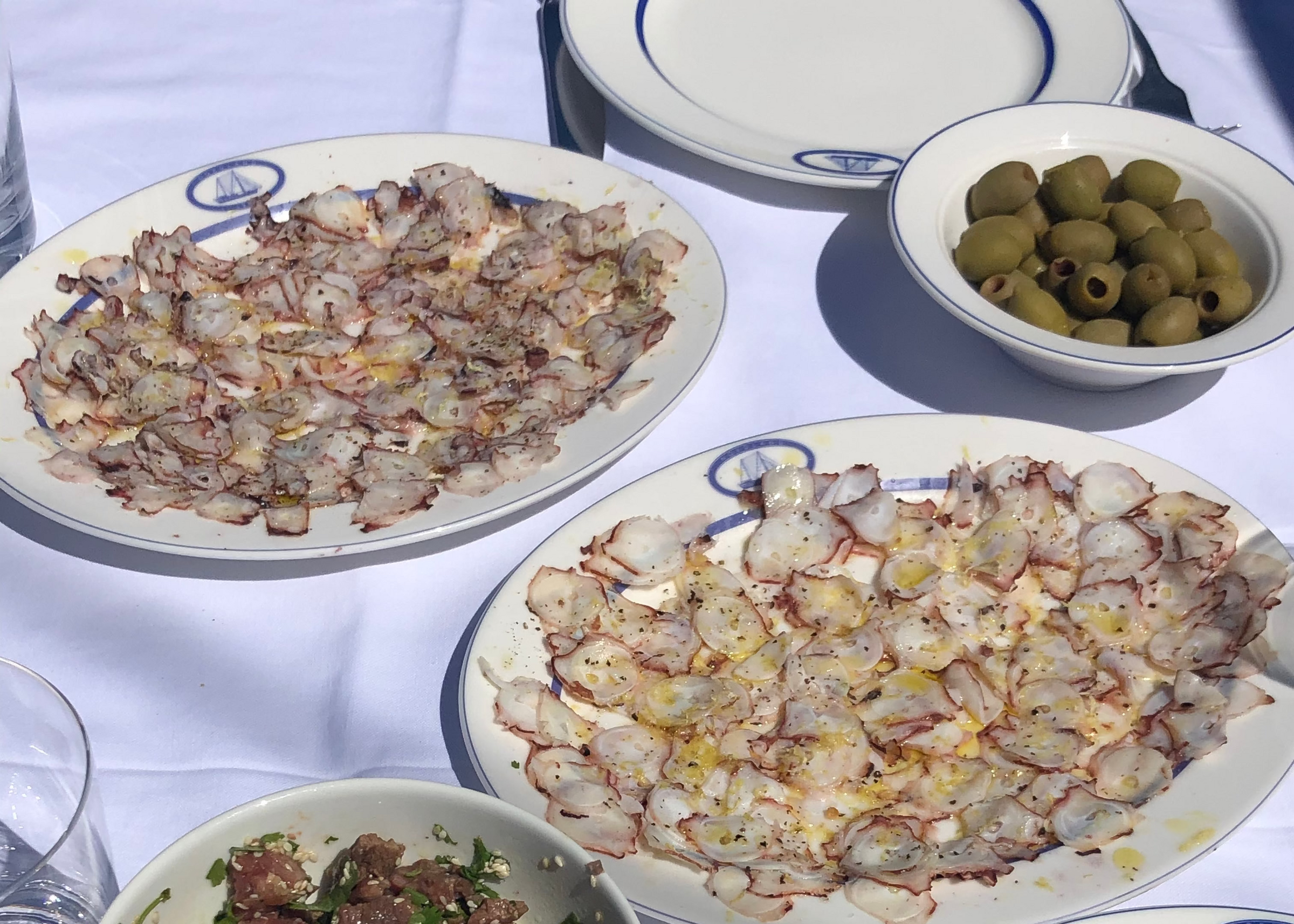 Plates of thinly sliced octopus, a bowl of green olives, and a bowl of marinated beef tartare with herbs are arranged on a white tablecloth with empty plates and glasses.