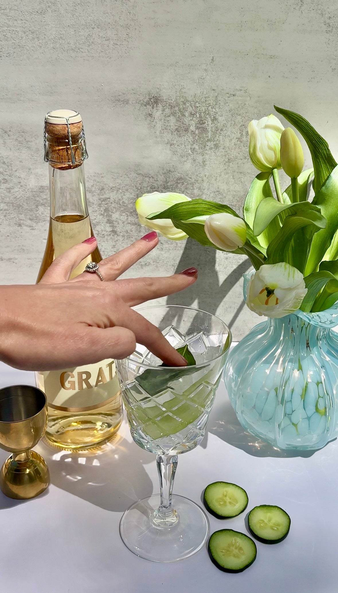 A hand with painted nails places a cucumber slice into a crystal glass with a drink. Nearby are cucumber slices, a gold cup, a bottle of wine, and a blue vase with white tulips against a light background.