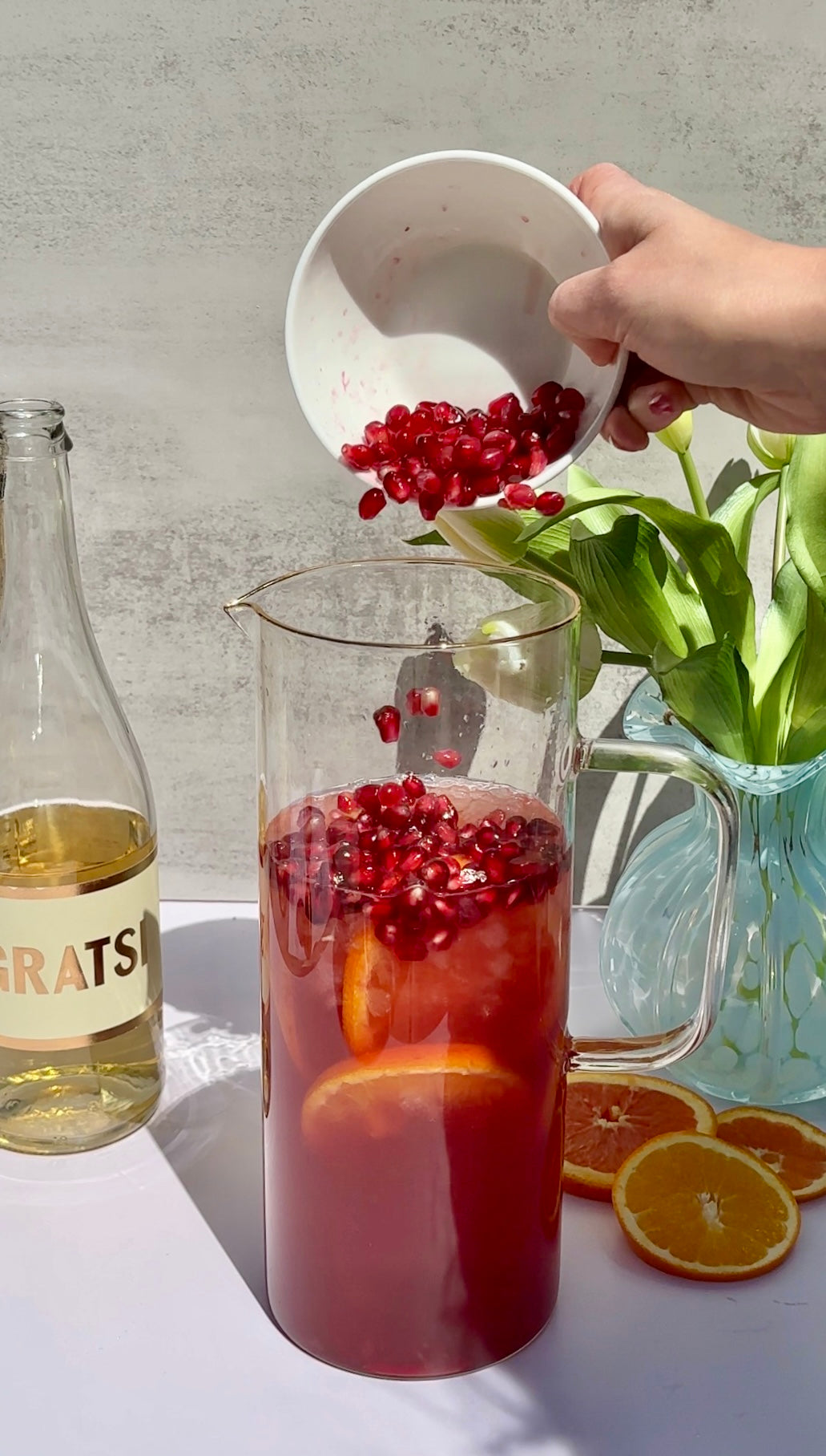A hand pours pomegranate seeds into a glass pitcher filled with a red drink and orange slices. Beside the pitcher are a bottle labeled GRATS, a clear vase with green leaves, and orange slices on a white surface.