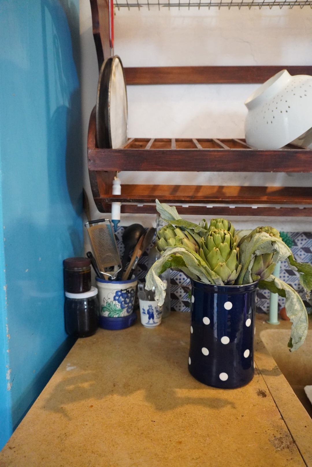 A bunch of artichokes in a blue polka dot mug sits on a kitchen counter, with utensils, a grater, jars, and a colander on a wooden rack in the background.