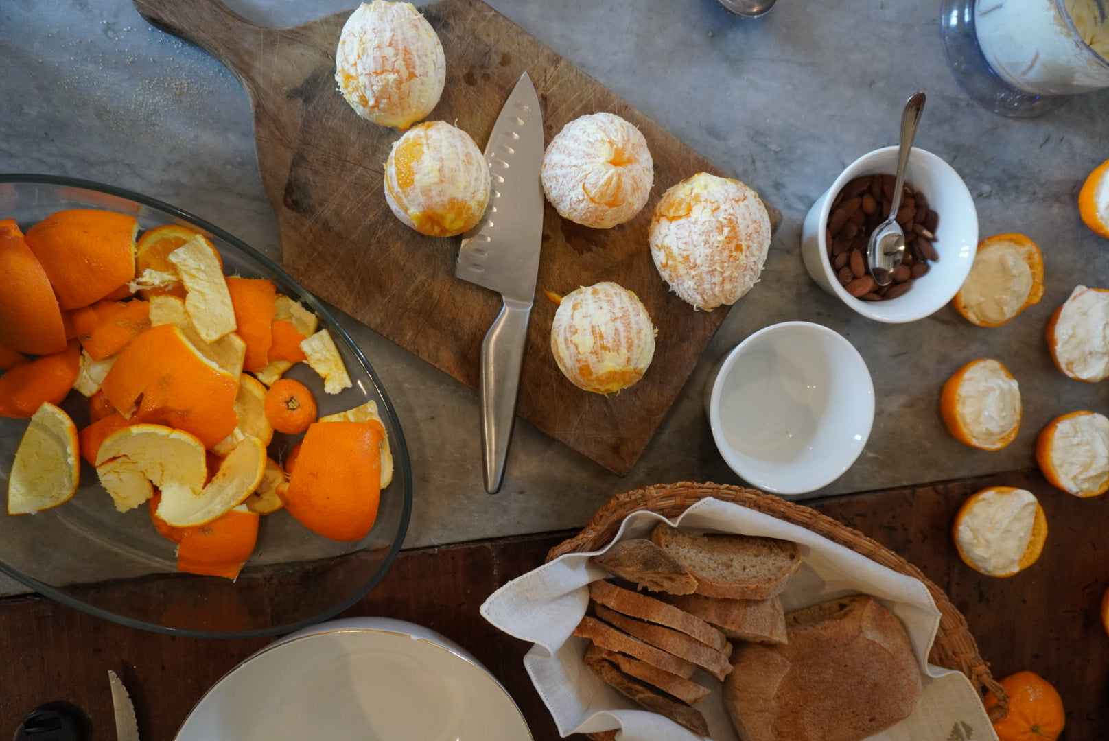 Peeled oranges, a knife, and orange peels sit on a cutting board. Nearby are bowls with bread slices, almonds, and empty orange halves on a marble and wood surface.