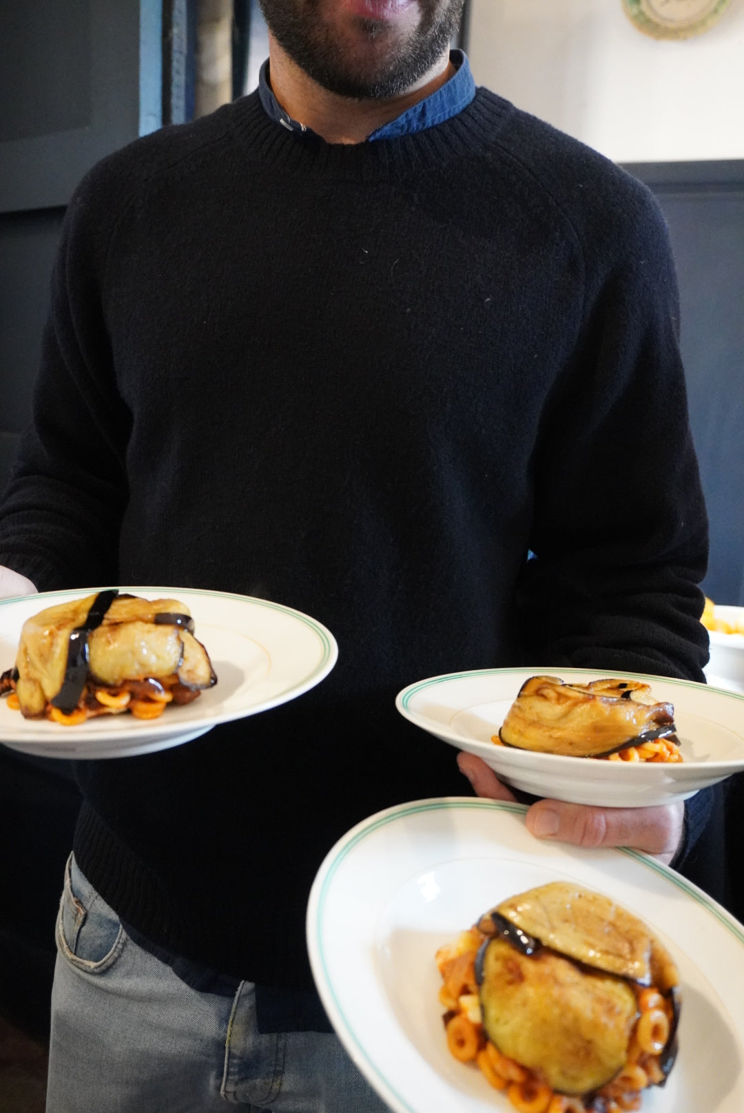 A person in a dark sweater holds three plates, each with a pastry topped with dark sauce and pasta underneath. Only the lower part of the persons face is visible.