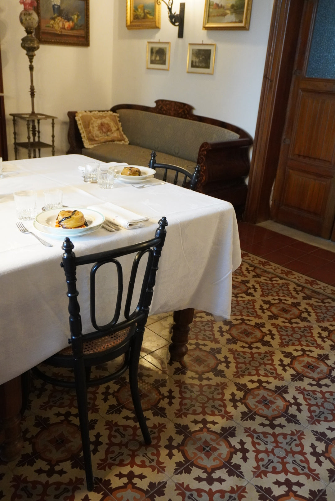 A dining table with a white tablecloth is set for two with plates of food, glasses, and cutlery. A decorative tile floor, wooden door, vintage sofa, and framed paintings are visible in a cozy, classic room.