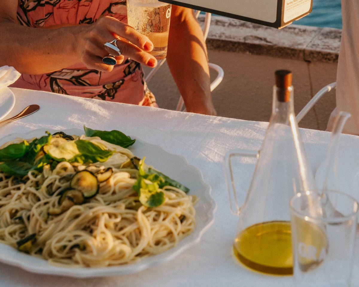 A person pours white wine from a Gratsi box into a glass at an outdoor table set with pasta, olive oil, and a woman in a pink floral top.