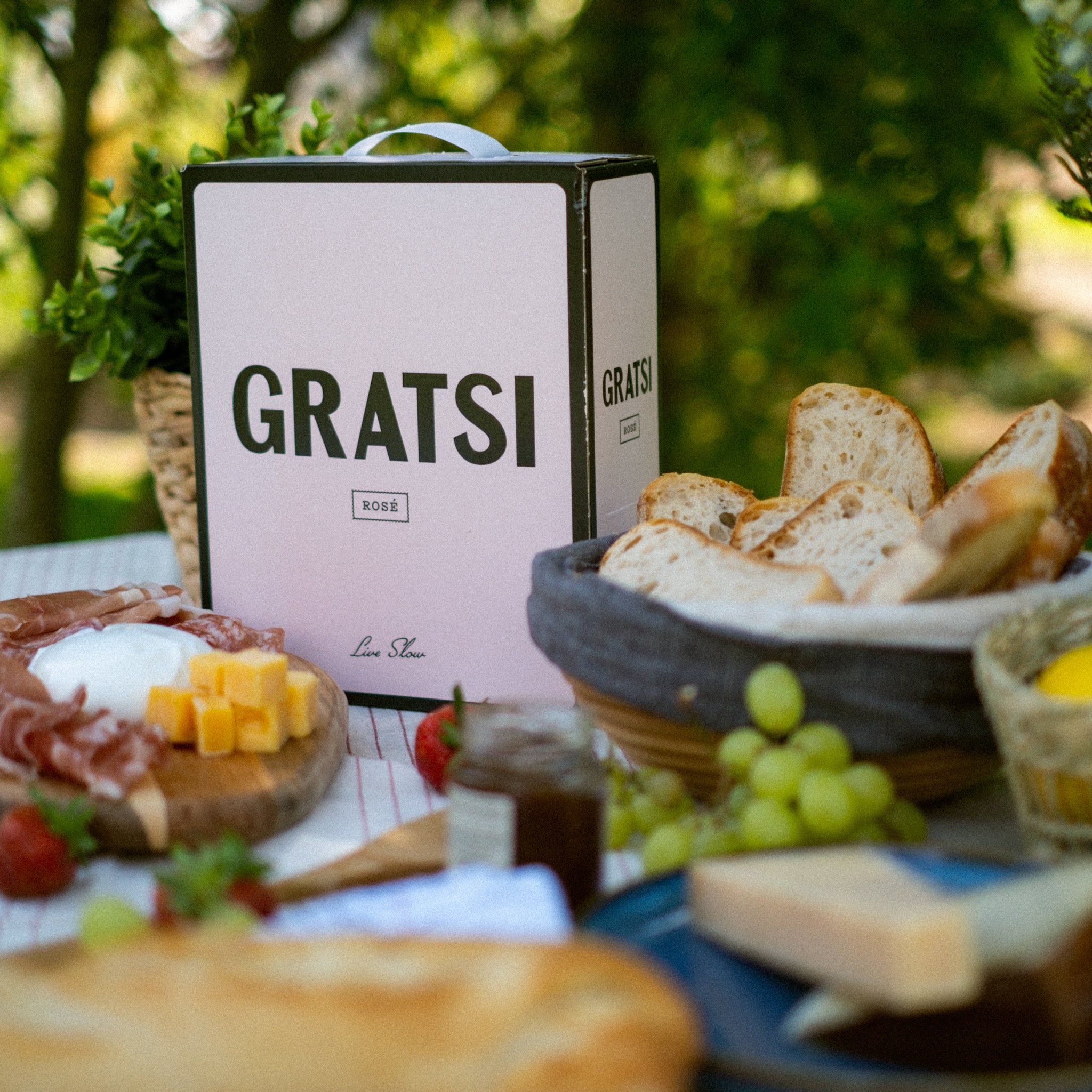 A picnic table with a box labeled “GRATSI Rosé,” baskets of sliced bread, grapes, cheese cubes, meat, strawberries, and jars of preserves, set outdoors with greenery in the background.