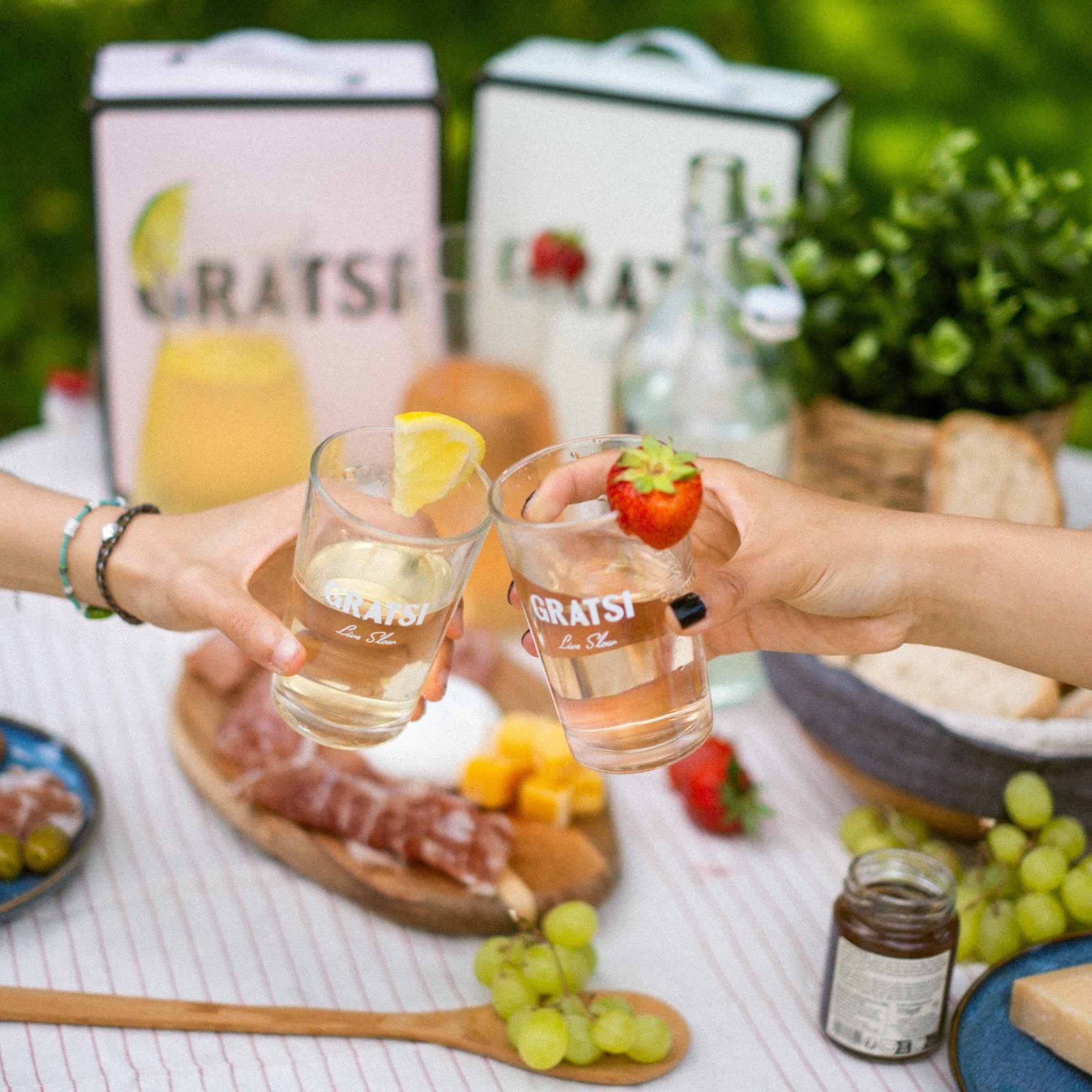Two people clink glasses with fruit garnishes over a picnic table set with grapes, cheese, bread, and drinks, with greenery and GRATS! beverage boxes in the background.