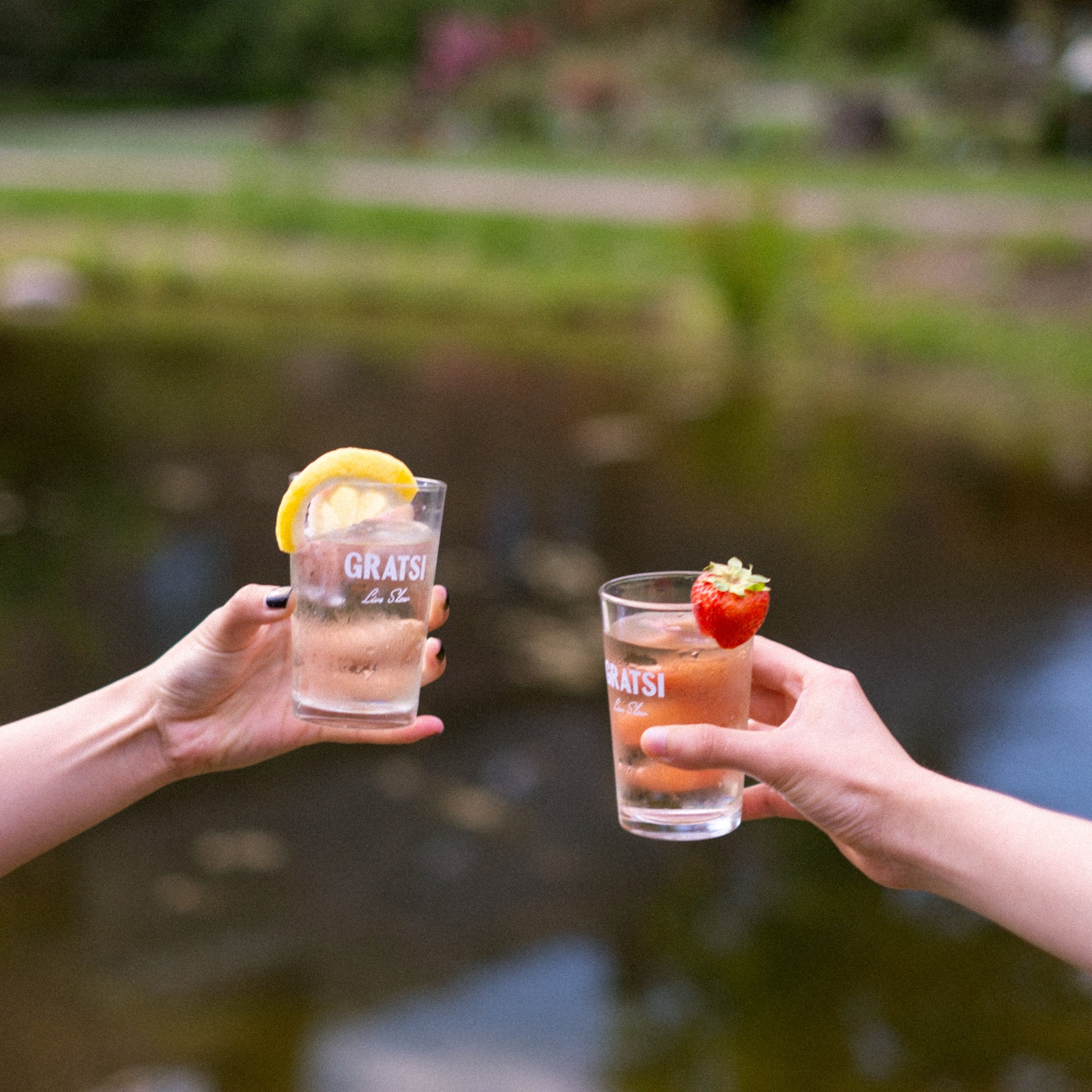 Two people hold up drinks in “GRATSI” glasses. One glass is garnished with a lemon slice; the other with a strawberry. A blurred pond and greenery appear in the background, suggesting an outdoor setting.