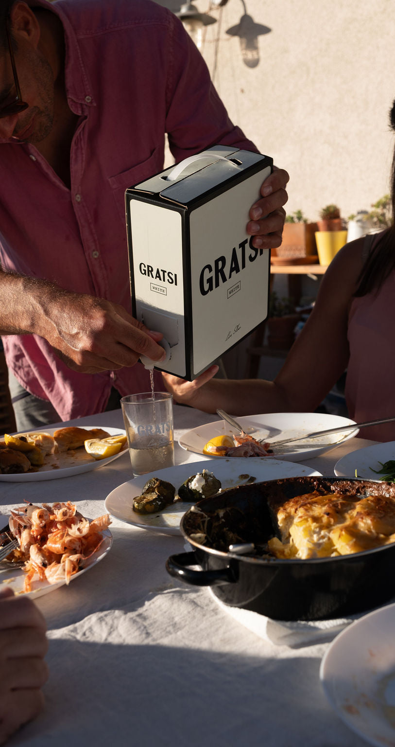 A person in a pink shirt pours wine from a GRATS! box into a glass at a sunlit outdoor table set with various dishes, including shrimp, grape leaves, and a baked casserole.