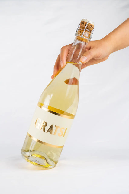 A hand holds a clear bottle of Gratsi Sparkling White with a cork top against a plain white background.