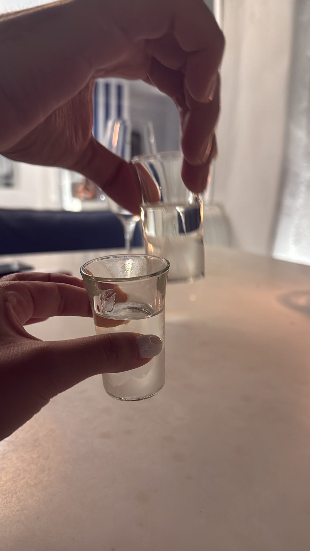Two hands each hold a shot glass filled with a clear liquid, reaching toward each other over a white table in a softly lit setting.