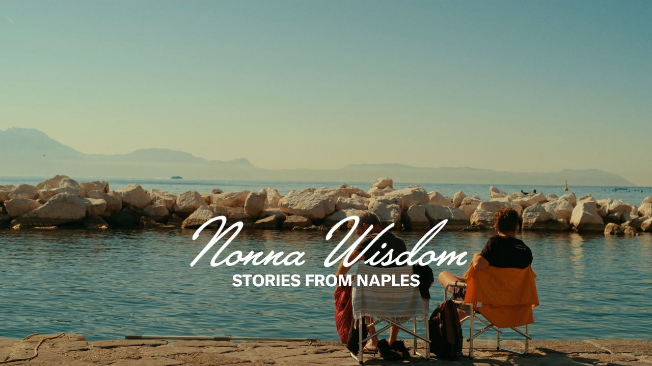Two people sit on chairs by the water facing rocky shore and distant mountains under a clear sky. Text reads: Nonna Wisdom - Stories from Naples.