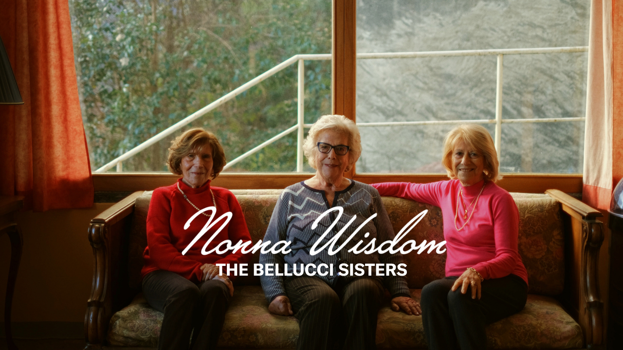 Three elderly women sit on a sofa in a cozy room with large windows. Text over the image reads: “Nonna Wisdom, The Bellucci Sisters.” The women are smiling and appear relaxed and content.
