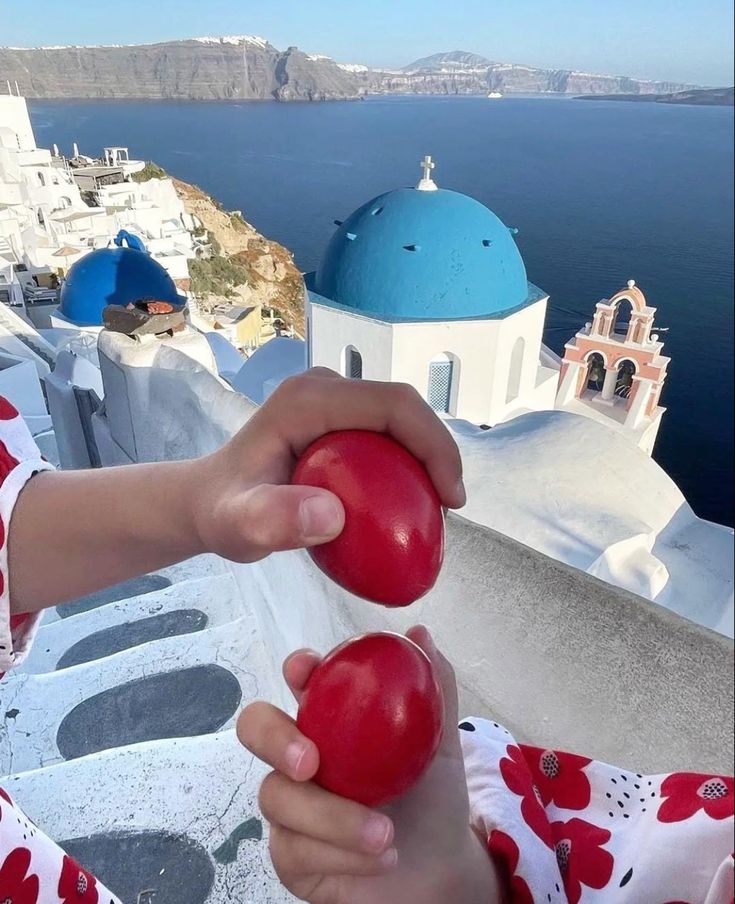 Two hands holding and tapping red Easter eggs in front of the iconic blue-domed church and white buildings of Santorini, Greece, overlooking the sea on a sunny day.