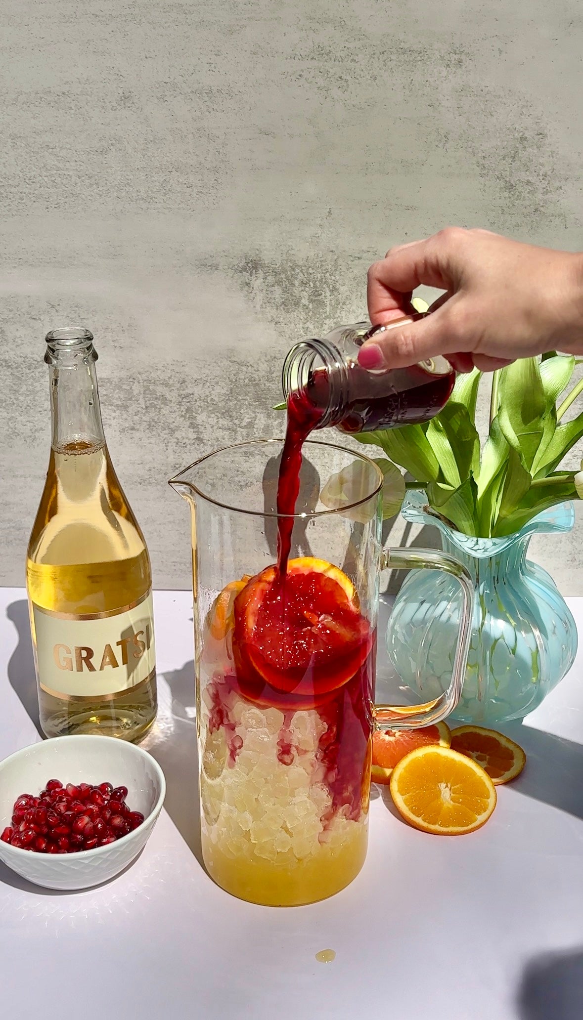 A hand pours red juice from a small bottle into a pitcher filled with ice and orange slices. Nearby are a bottle of sparkling wine, a bowl of pomegranate seeds, orange slices, and a vase of green tulips.
