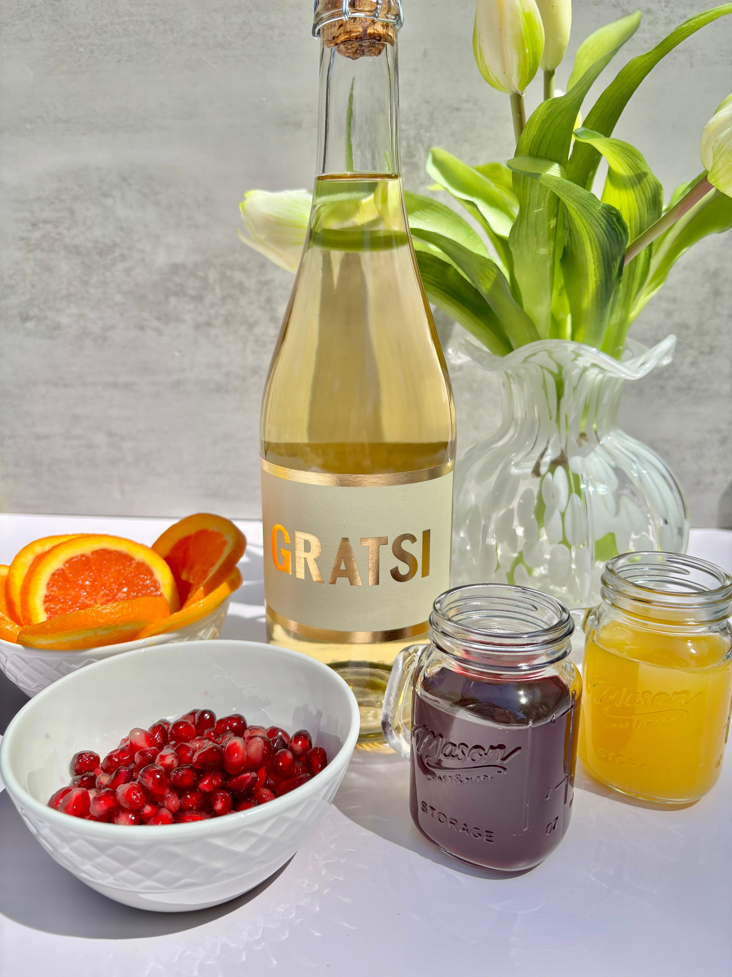 A bottle of Gratsi wine stands on a table with sliced oranges, a bowl of pomegranate seeds, two mason jars of red and yellow juice, and a vase of white tulips in the background.