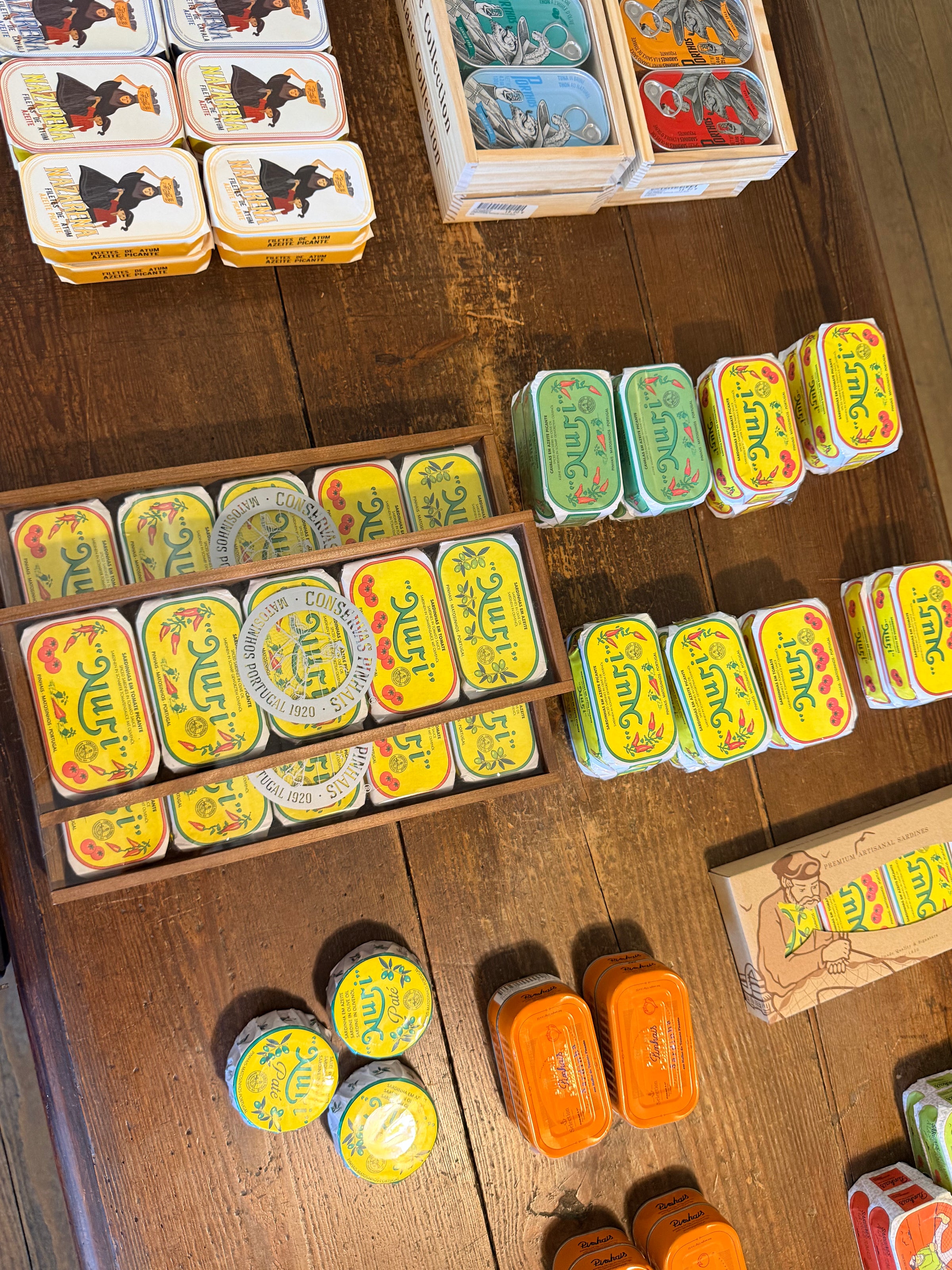 Colorful tins of canned fish, mainly yellow and orange, are neatly arranged on a wooden table. Some round tins and rectangular boxes are stacked together, displaying various artwork and branding.