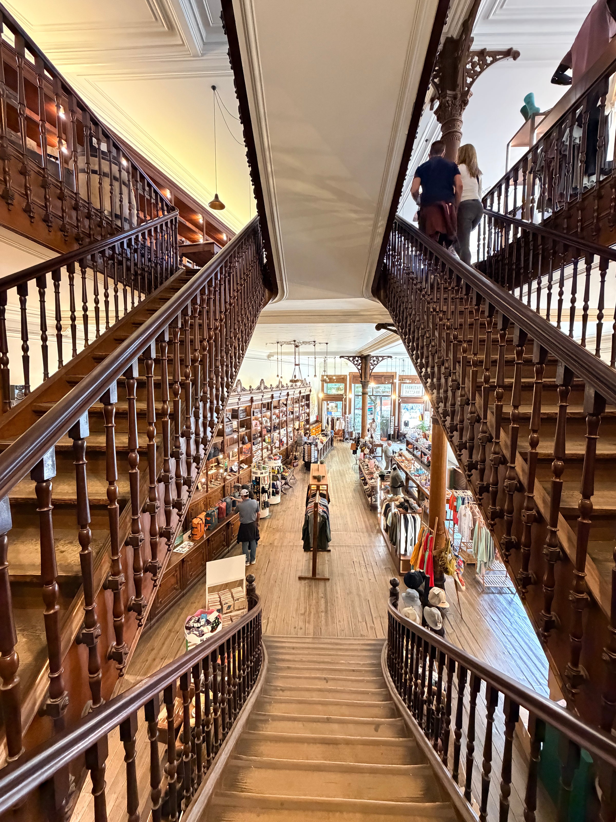 A view from the top of a grand wooden staircase in a large, vintage-style clothing store, with shelves, racks of clothes, and shoppers visible below on the main floor.