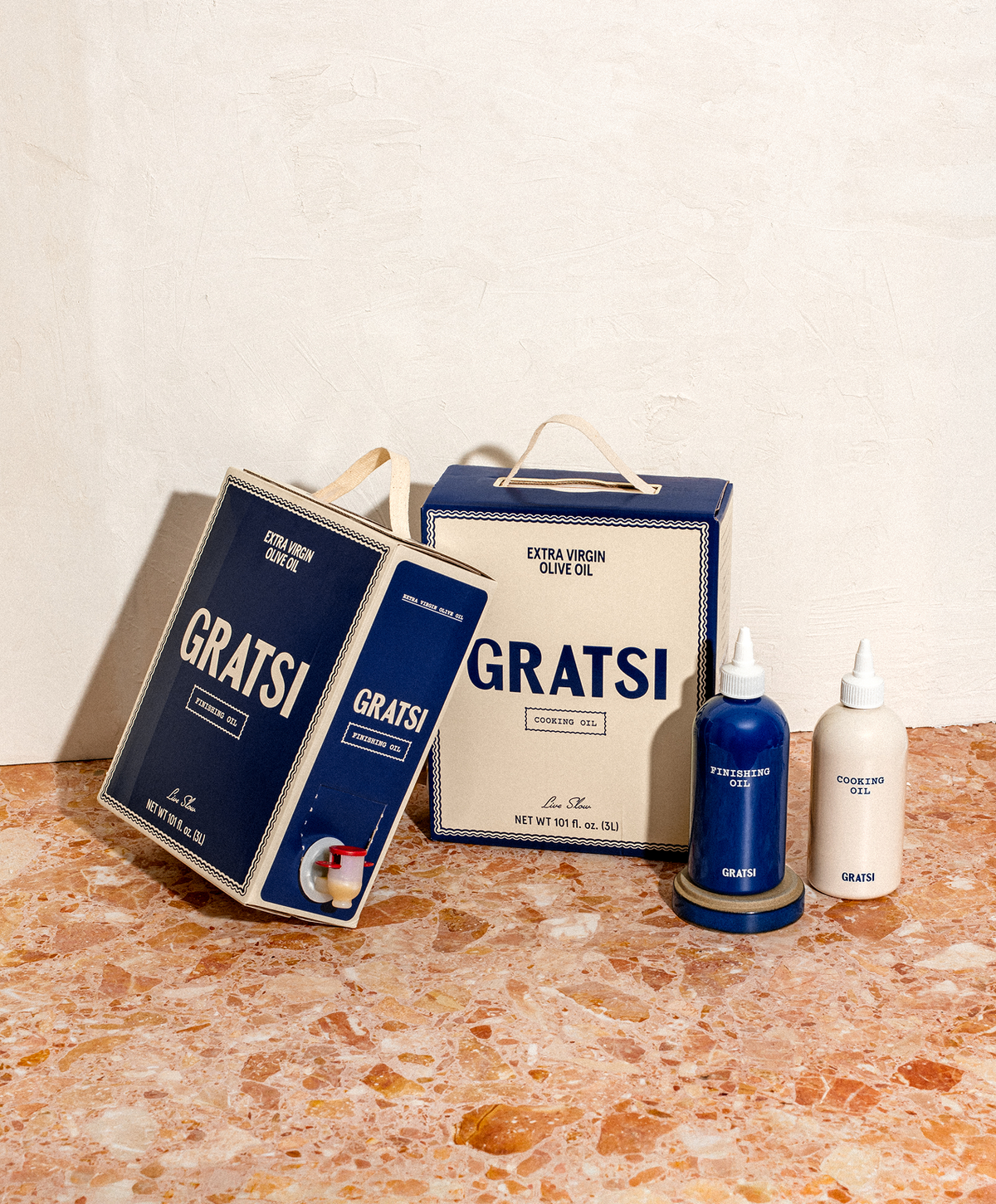 Two boxes of Gratsi extra virgin olive oil and two squeeze bottles of finishing and cooking oil on a marble surface.