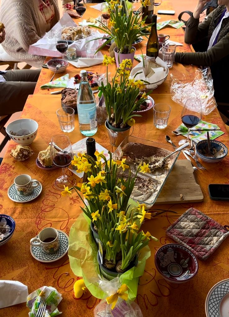 A table set with coffee cups, plates, wine glasses, water, food remnants, and yellow daffodils in pots. Several people sit around the orange tablecloth, suggesting a finished meal or gathering.