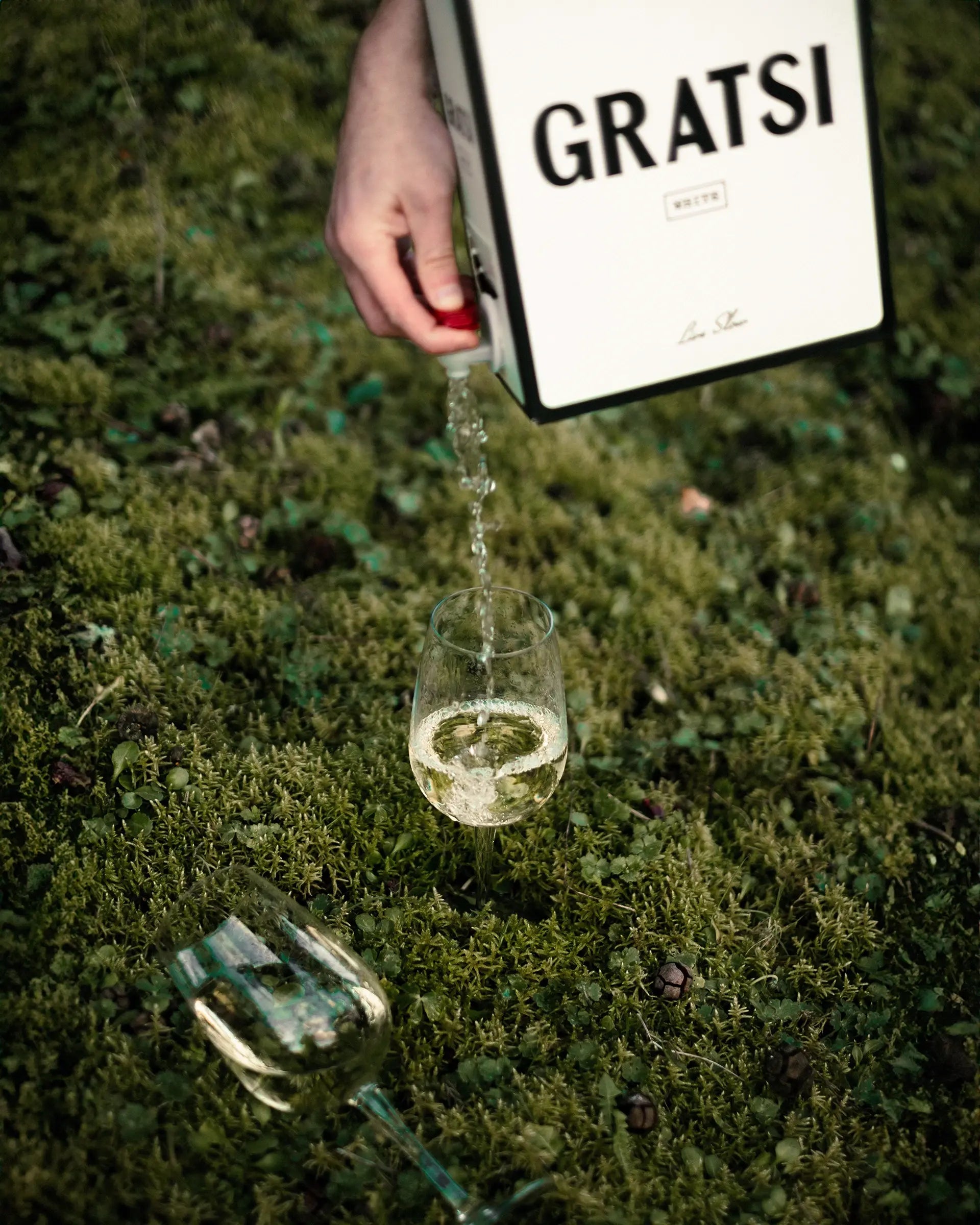 A hand pours white wine from a large box labeled GRATSI into a wine glass on grass, with another empty wine glass lying nearby.