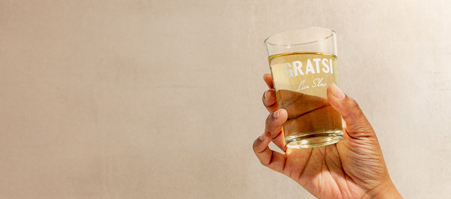 A hand holding a glass filled with a light yellow beverage. The glass has the words GRATSI Live Slow printed on it, and the background is a plain, light beige color.