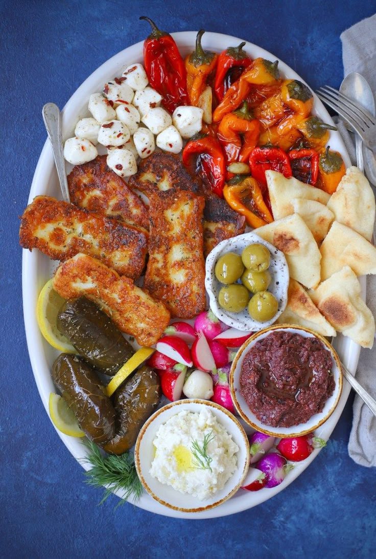 A platter with grilled halloumi, stuffed grape leaves, marinated mozzarella, roasted peppers, olives, pita bread, radishes, and bowls of hummus and tapenade, arranged on a white dish against a blue background.